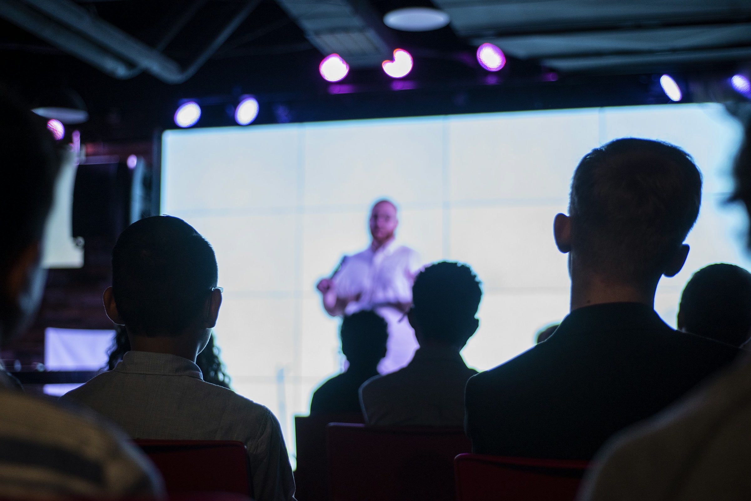 Audience seen from behind in a darkened room, watching a speaker in front of a bright projection screen