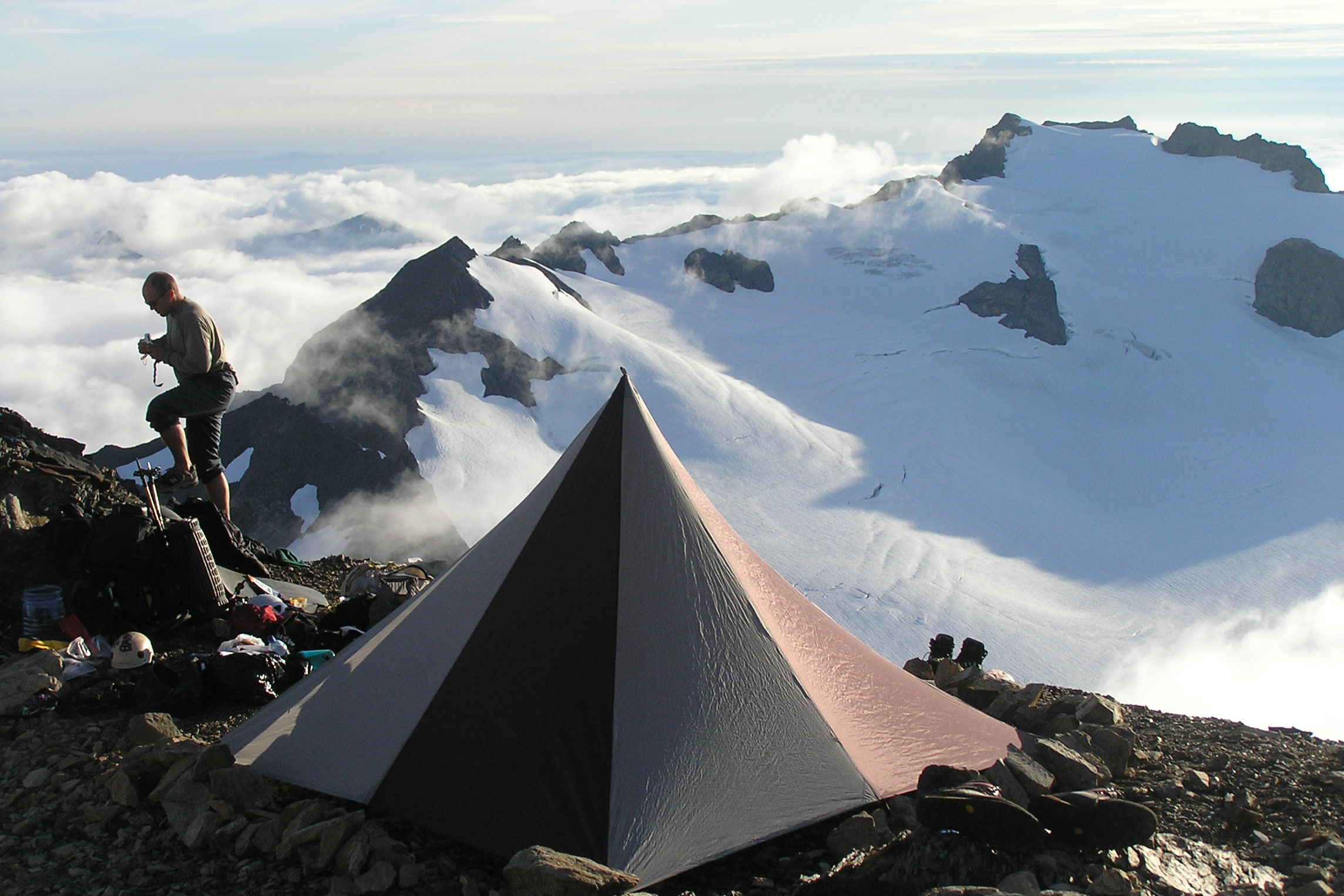 A backpacker climbing rocks beside a pyramid tent on a high ridge above a sea of clouds and a snowy peak