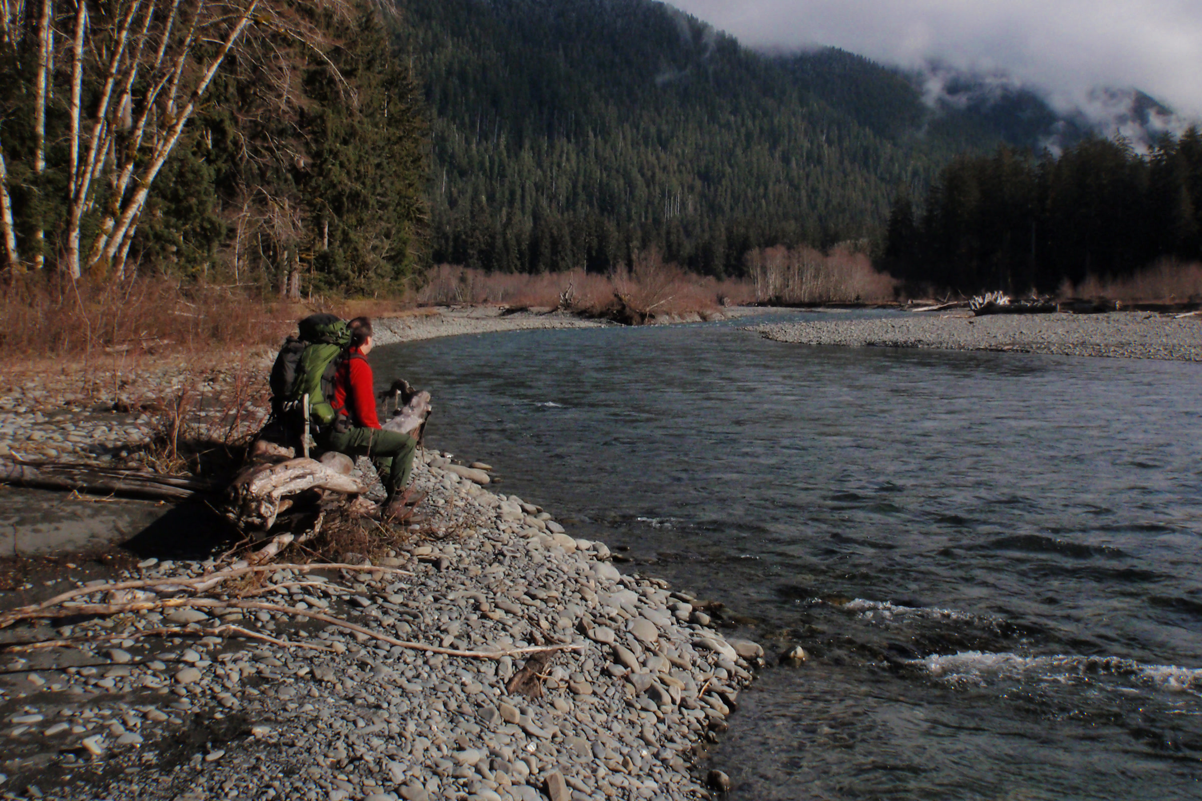 Two backpackers resting on a driftwood log on a gravel bar beside the Hoh River, with forested hills and low clouds behind