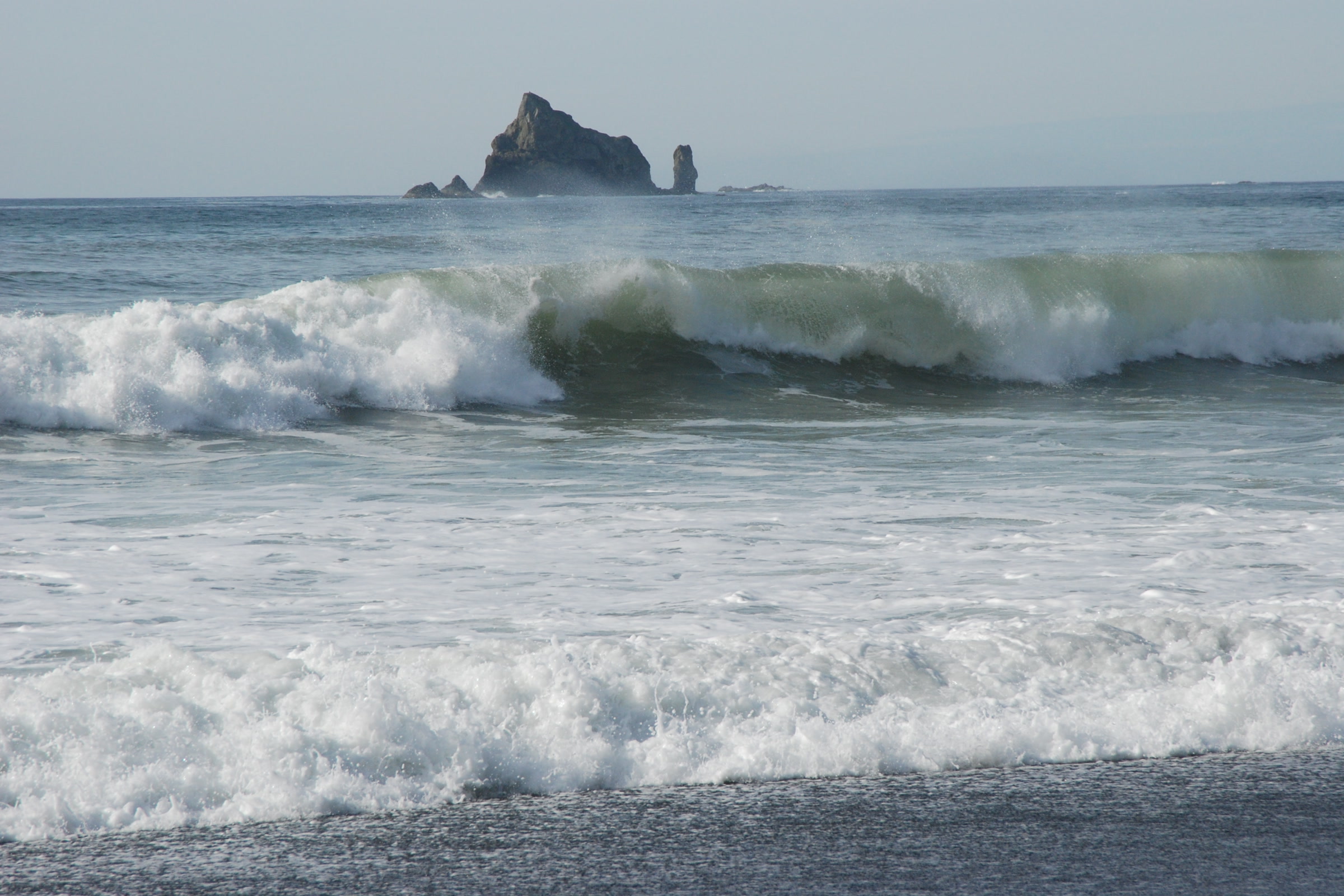 Big green waves breaking toward the steep shore at Rialto Beach, with a rocky sea stack on the horizon