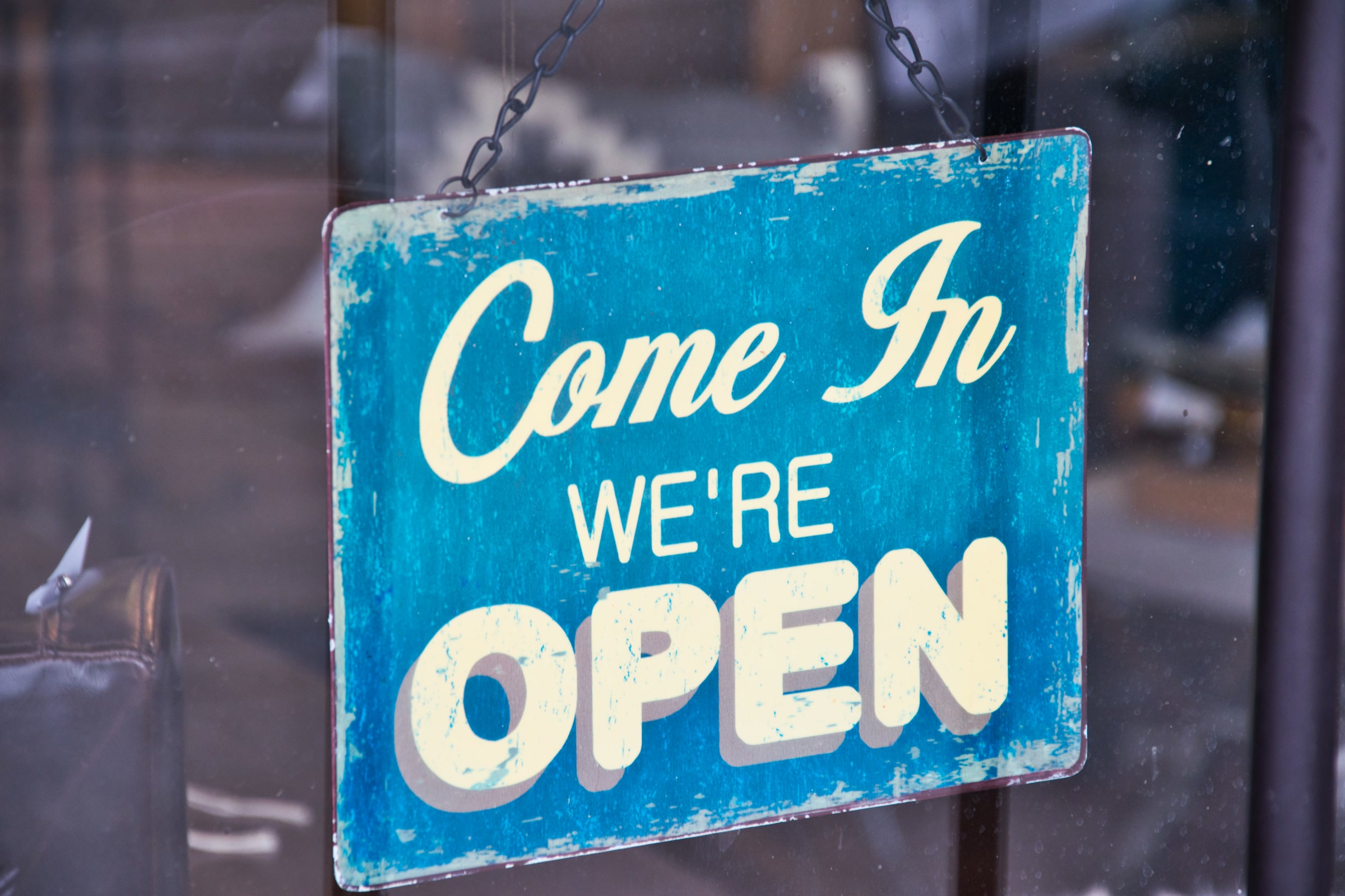 A weathered blue and cream Come In We're Open sign hanging in a shop's glass door