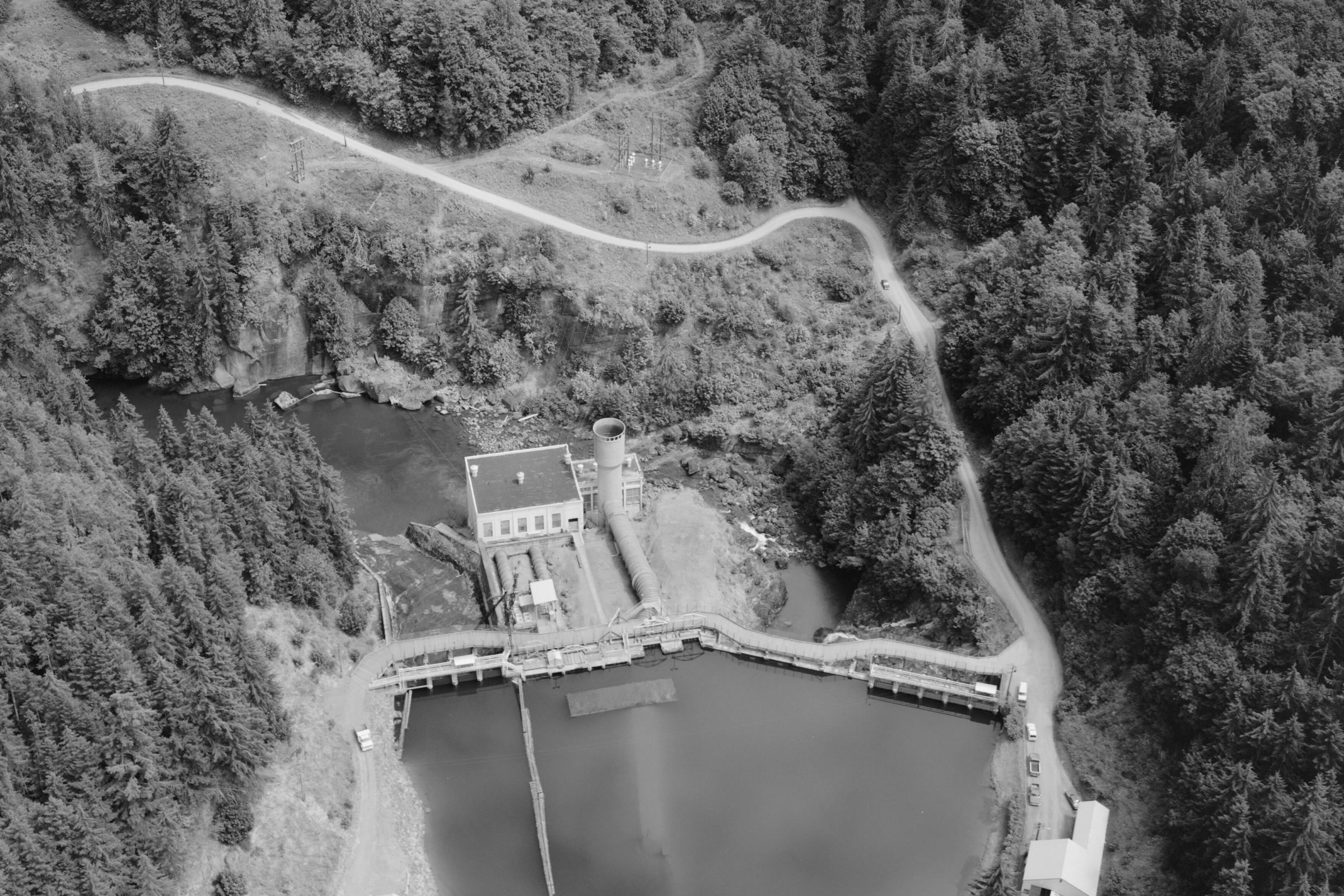 Black and white aerial view of the Elwha Dam and powerhouse holding back Lake Aldwell, with a road curving through the forest above