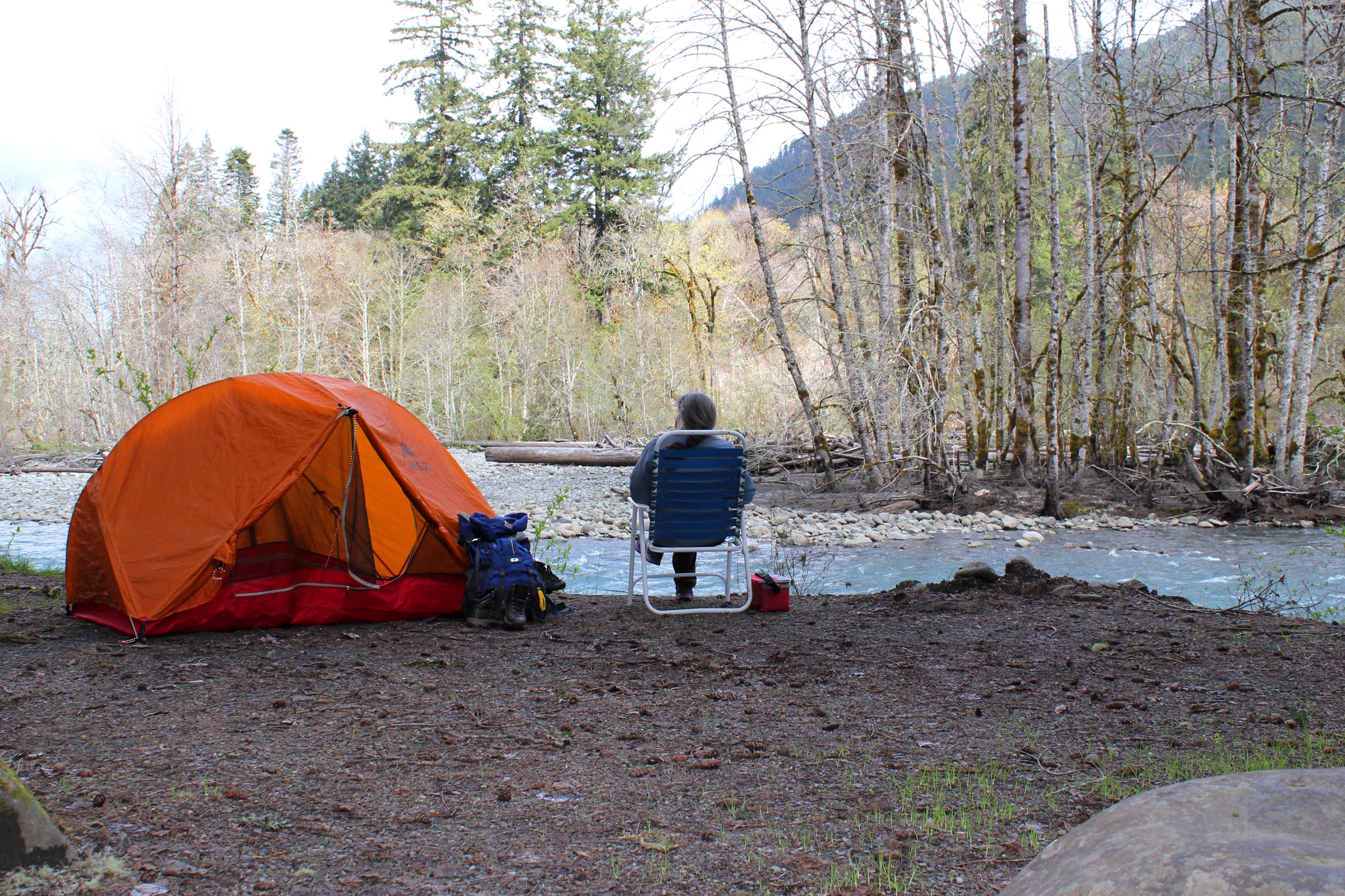 An orange tent and a camp chair on a gravel campsite beside the Elwha River, with alder and conifer forest on the far bank