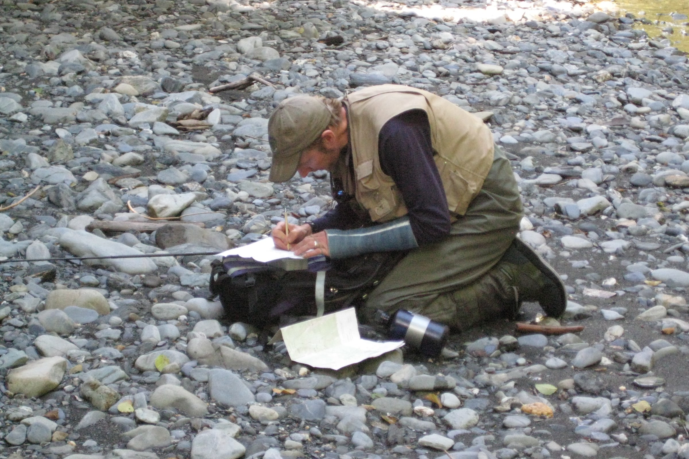 An Olympic National Park fisheries biologist in waders kneels on a gravel river bar writing survey notes beside his gear