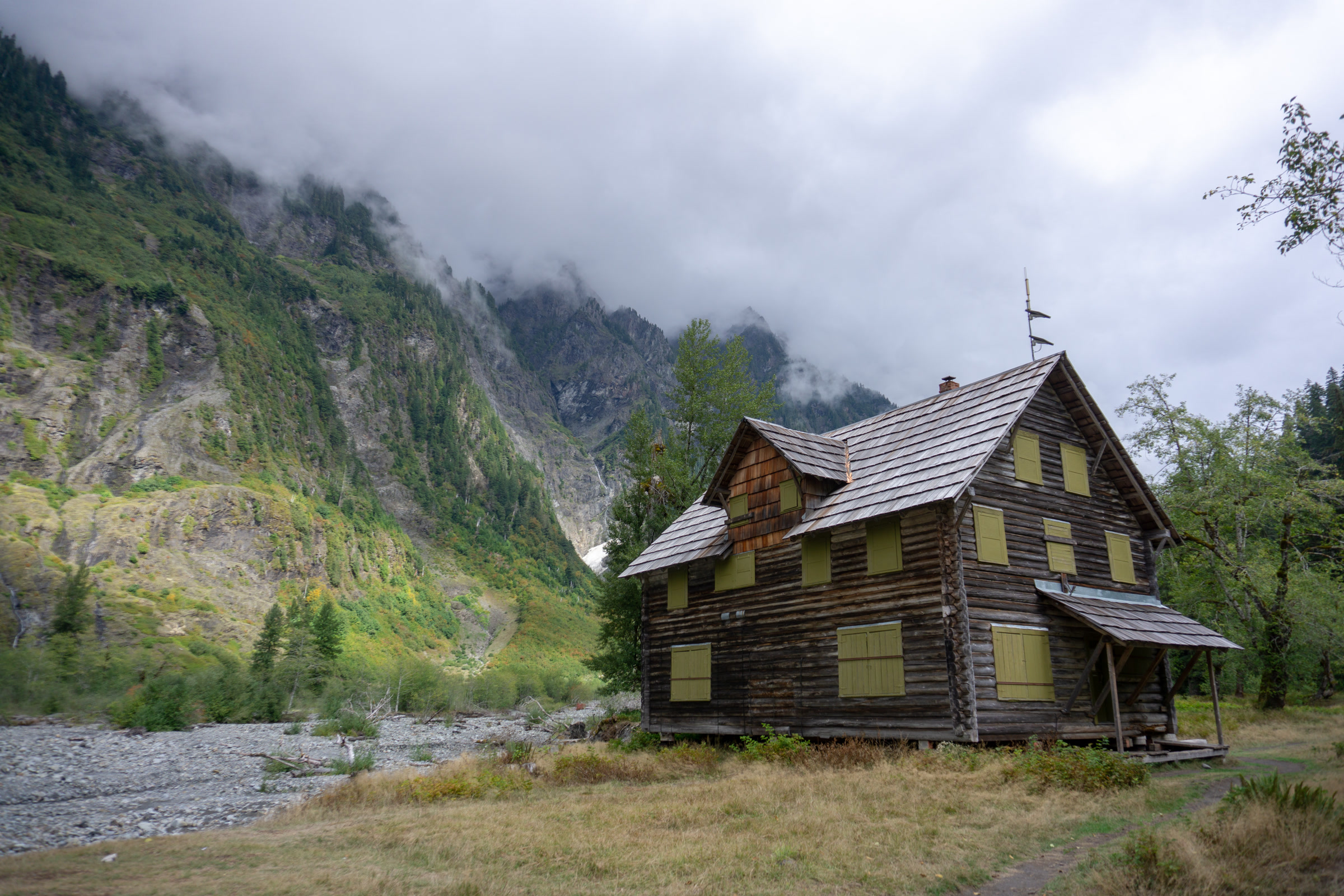 The three-story log Enchanted Valley Chalet with boarded yellow windows, standing on a grassy terrace below cloud-wrapped cliffs