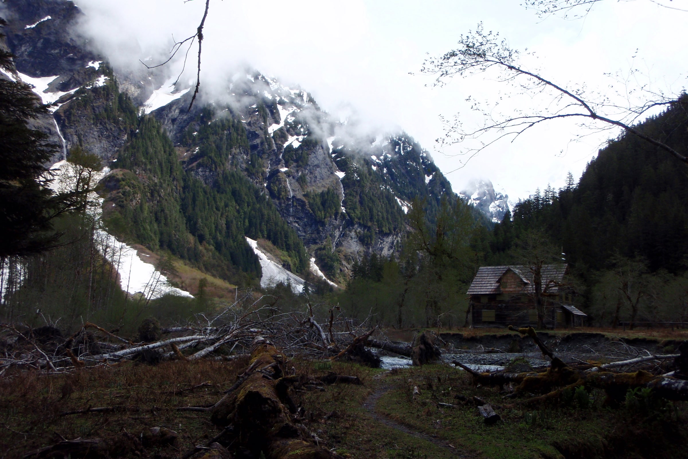 The Enchanted Valley Chalet small in the distance beyond a jumble of fallen logs and gravel, with snow-streaked cliffs rising behind