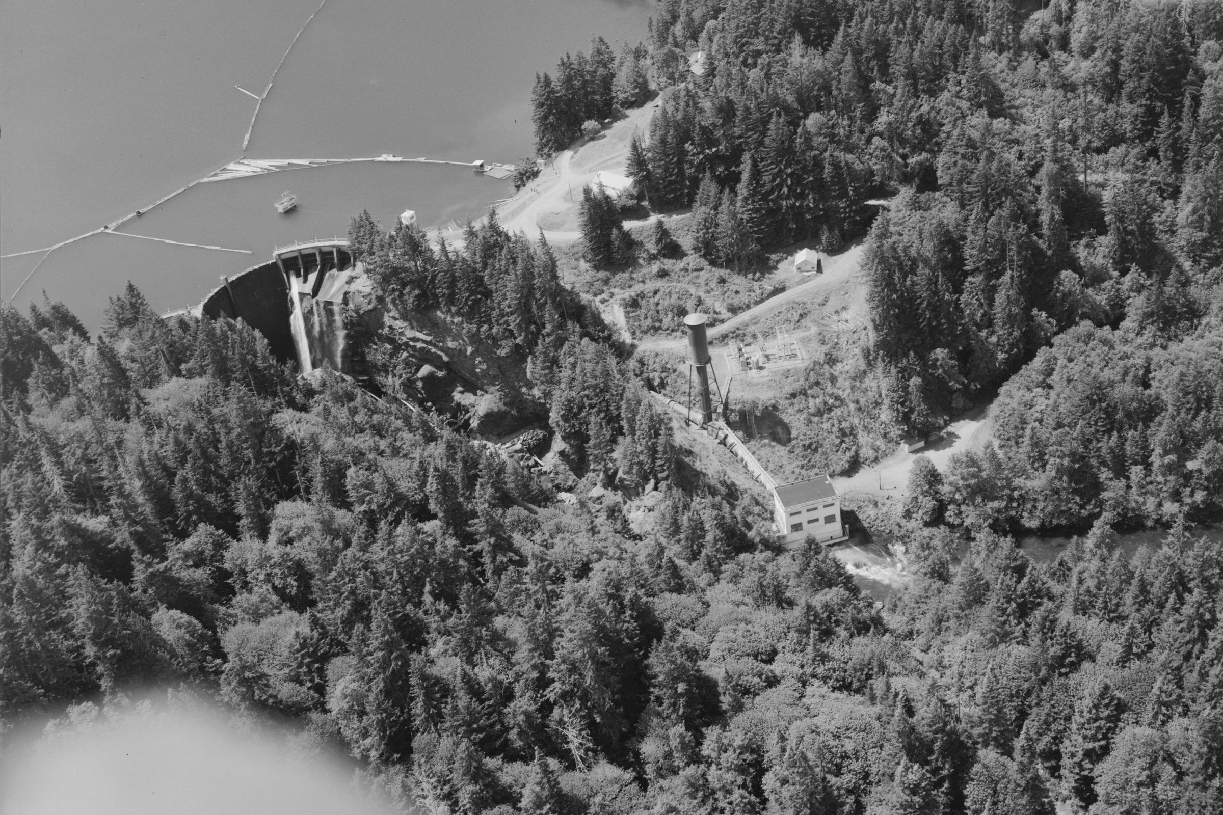 Black and white aerial view of the curved Glines Canyon Dam holding back Lake Mills, with its surge tank and powerhouse in the forest below