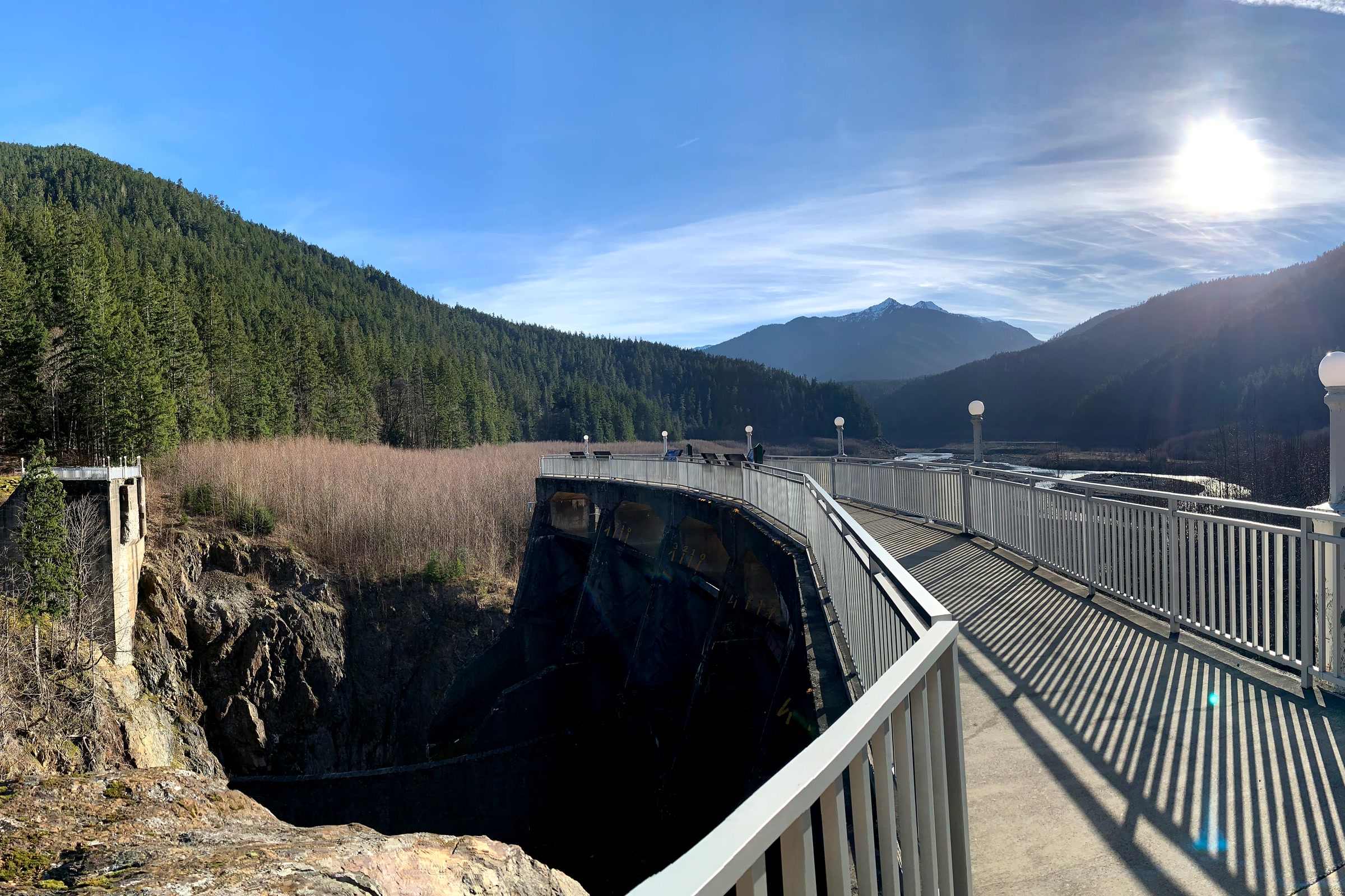 A railed walkway on top of the old Glines Canyon Dam spillway, looking up the forested Elwha valley under a bright winter sun