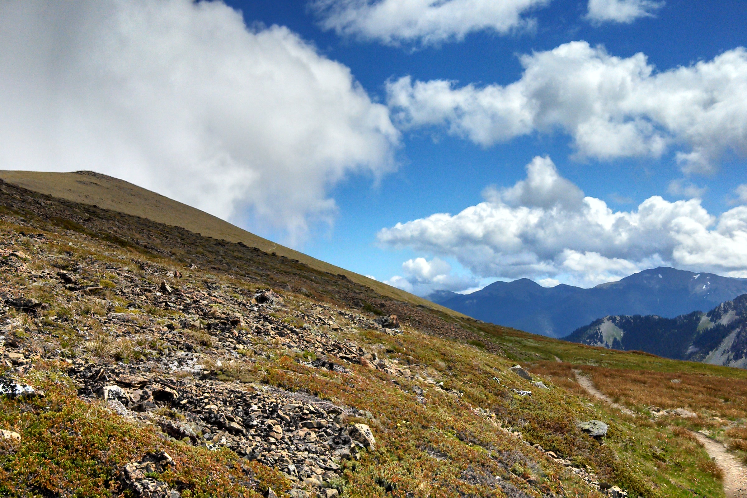 A narrow trail crossing a rocky, red tinged subalpine slope under big white clouds, looking east toward Deer Park