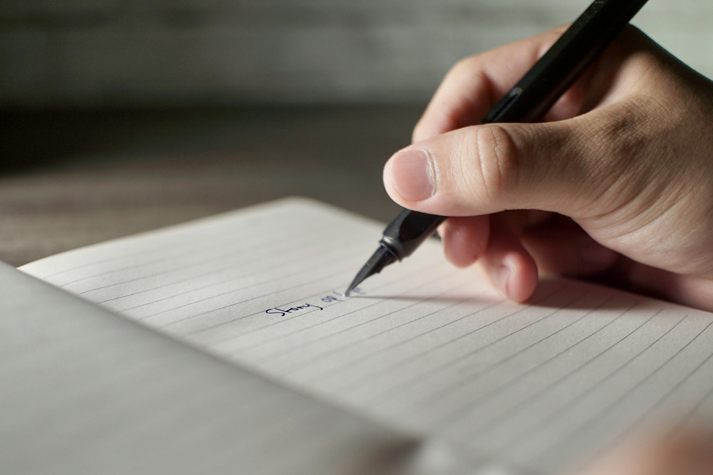 A hand writes with a black pen on a lined notebook page in soft light