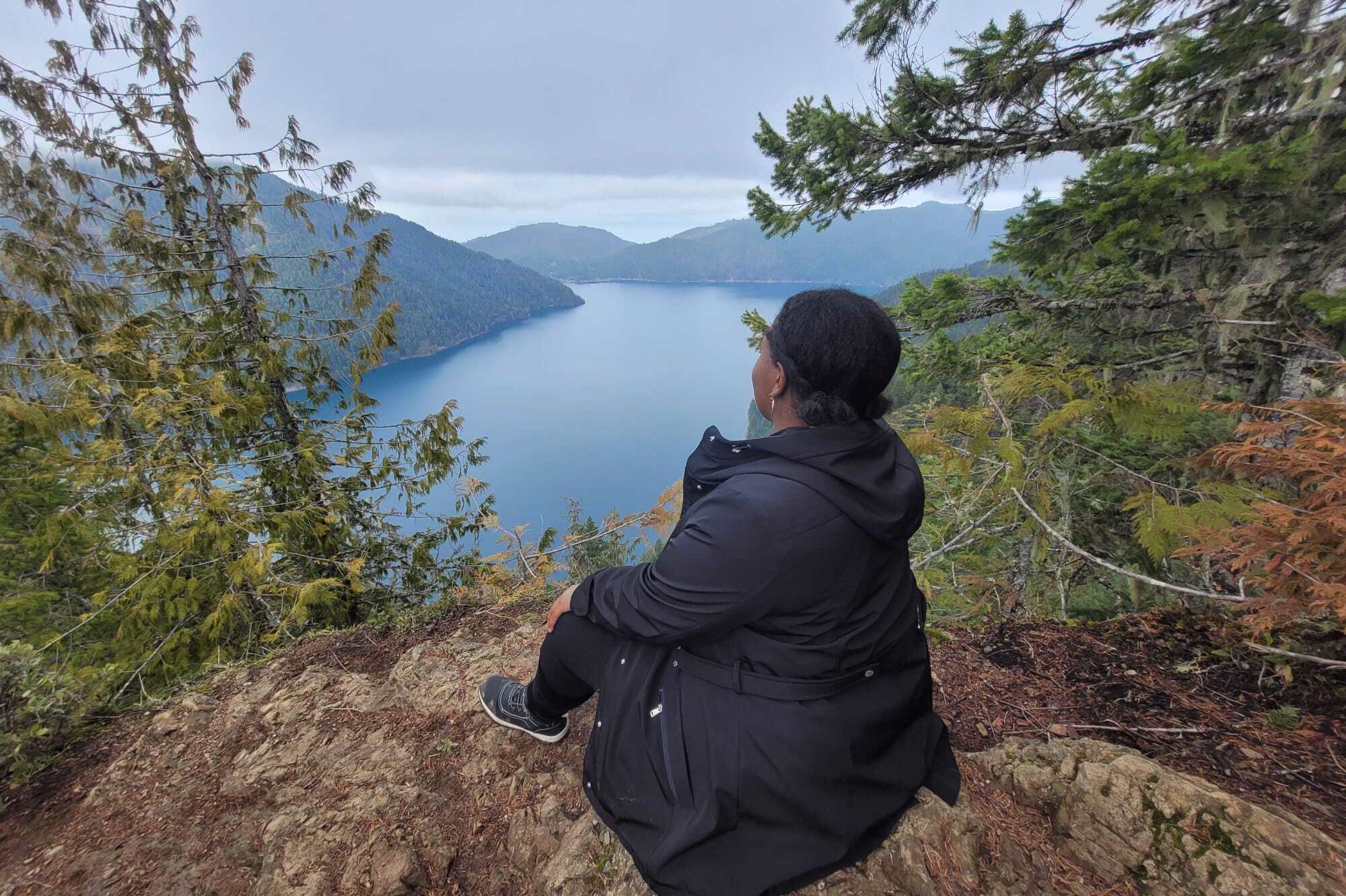 A hiker in a dark jacket sitting on bare rocky ground on Mount Storm King, looking out over the long blue sweep of Lake Crescent