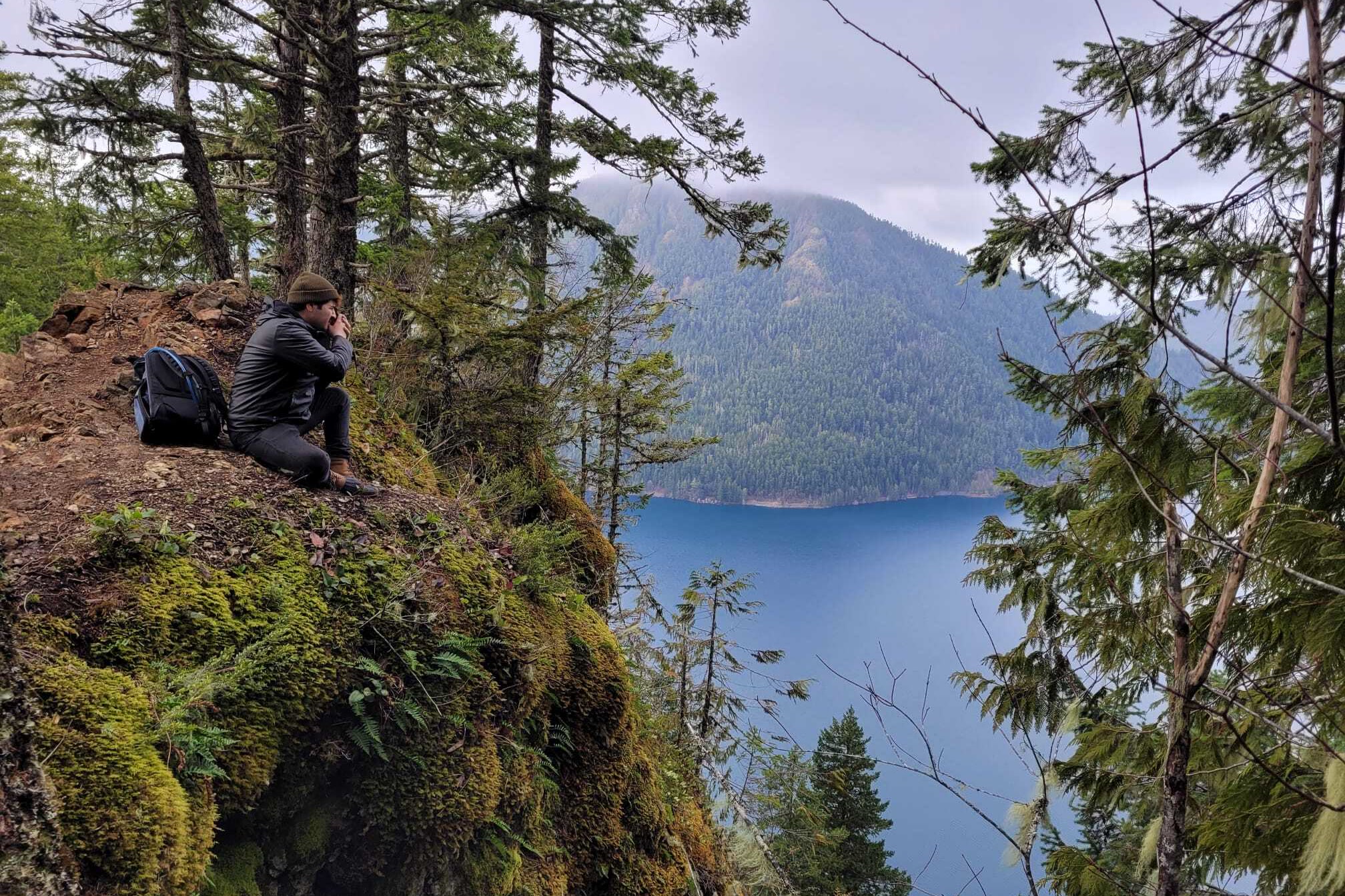 A hiker sitting on a mossy rock outcrop on Mount Storm King, photographing Lake Crescent far below between forested slopes