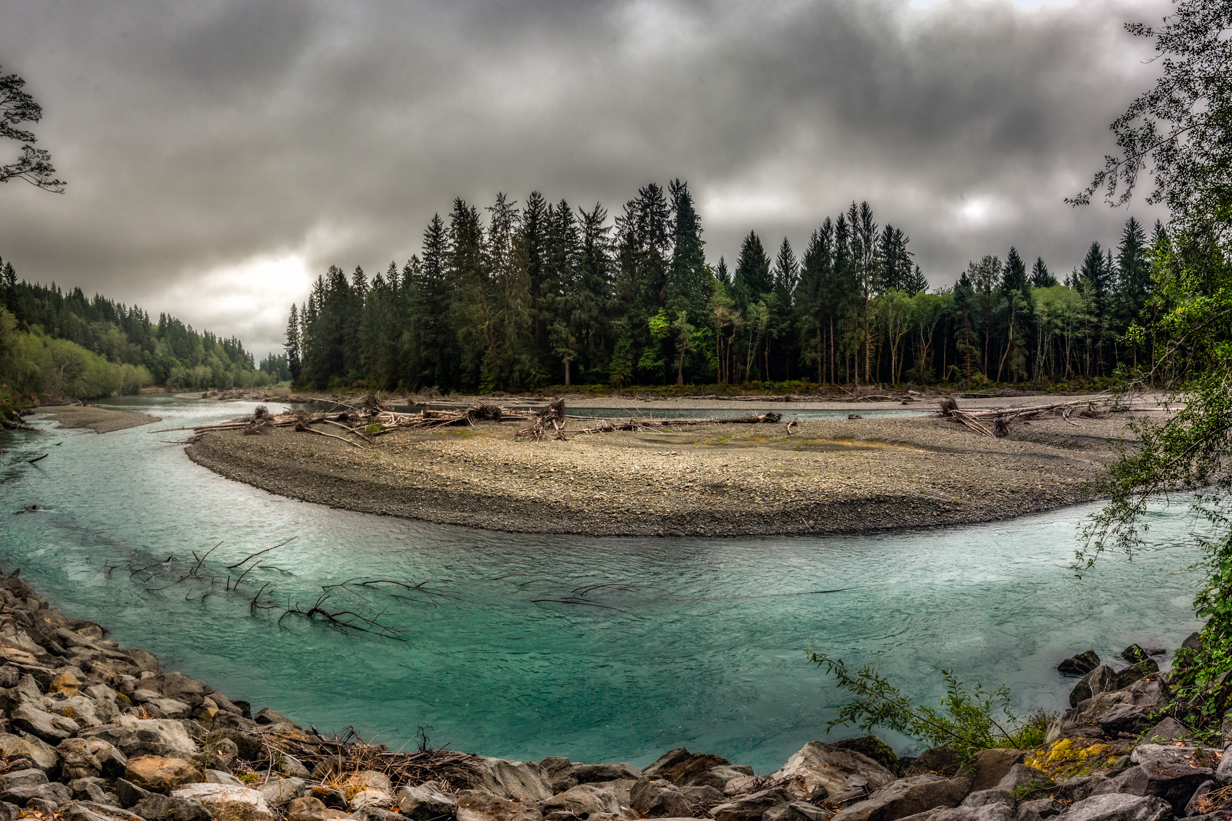 The blue-green Hoh River curving around a wide gravel bar lined with driftwood and tall conifers under grey skies