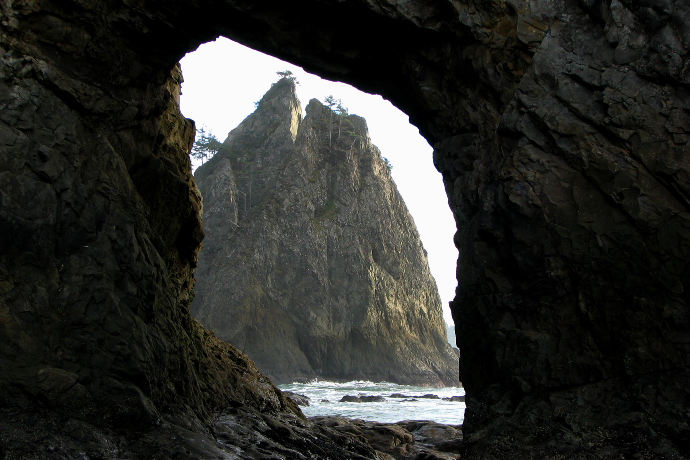 View through the dark rock arch of Hole-in-the-Wall north of Rialto Beach toward a tall sea stack and surf