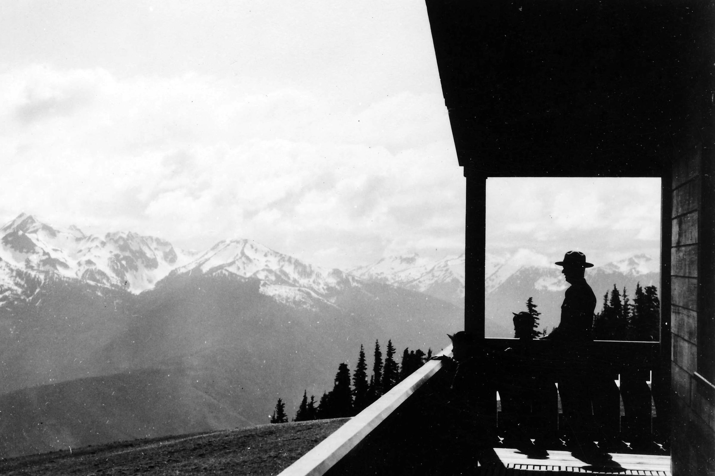 Black and white photo of a figure in a flat hat on a lodge porch looking out at snowy Olympic peaks