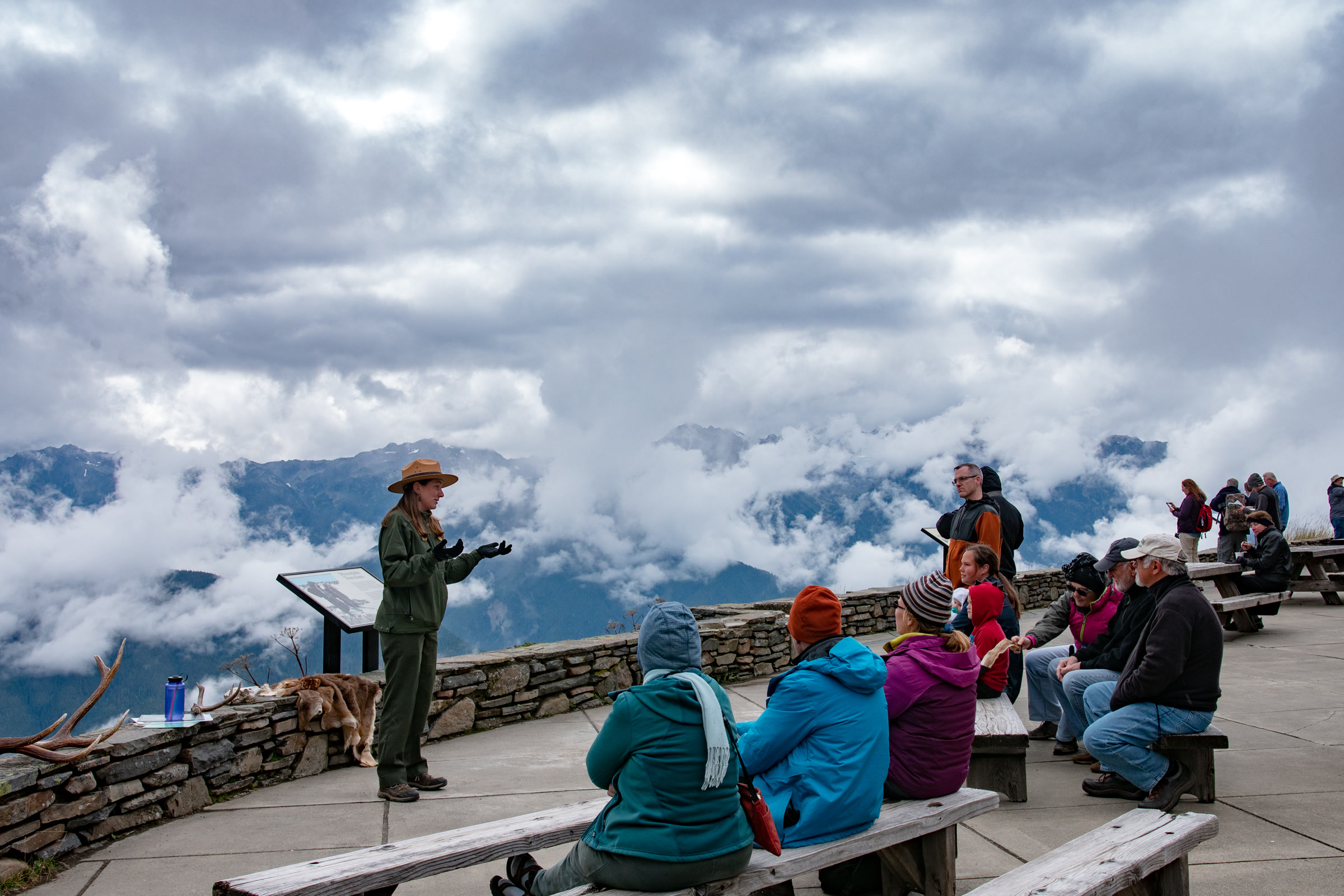 A uniformed park ranger gives a talk to visitors seated on benches on the Hurricane Ridge Visitor Center patio, with clouds over the mountains behind