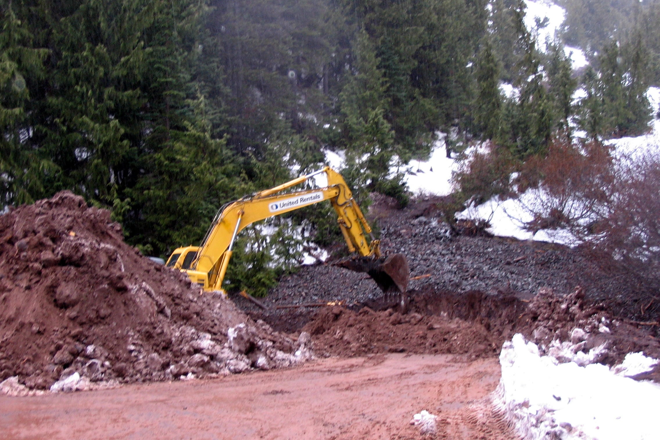 A yellow backhoe clears red earth and rock debris beside snowbanks at the Switchback Trailhead on Hurricane Ridge Road