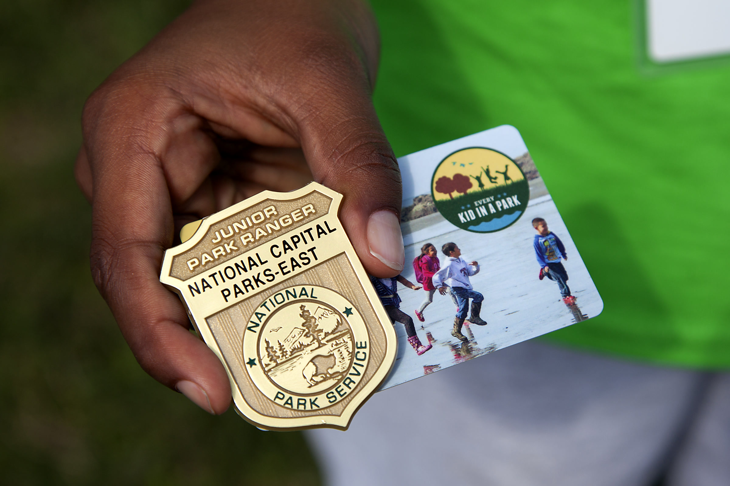 A child's hand holding a gold Junior Park Ranger badge next to a fourth-grade park pass