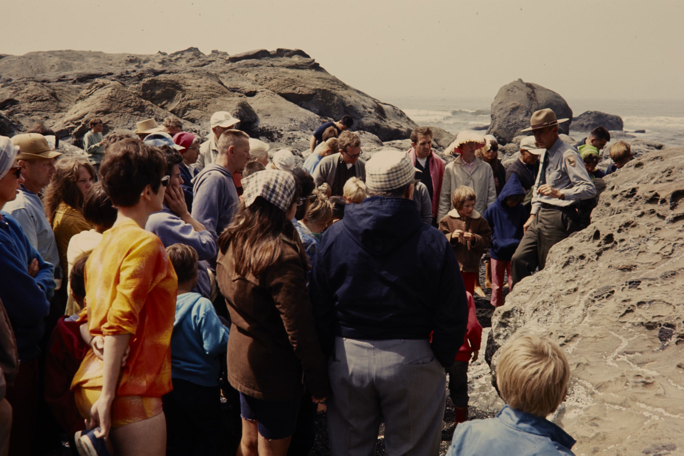 A uniformed park naturalist in a flat hat talks to a large crowd of visitors gathered among sandstone rocks on a Kalaloch tidepool walk