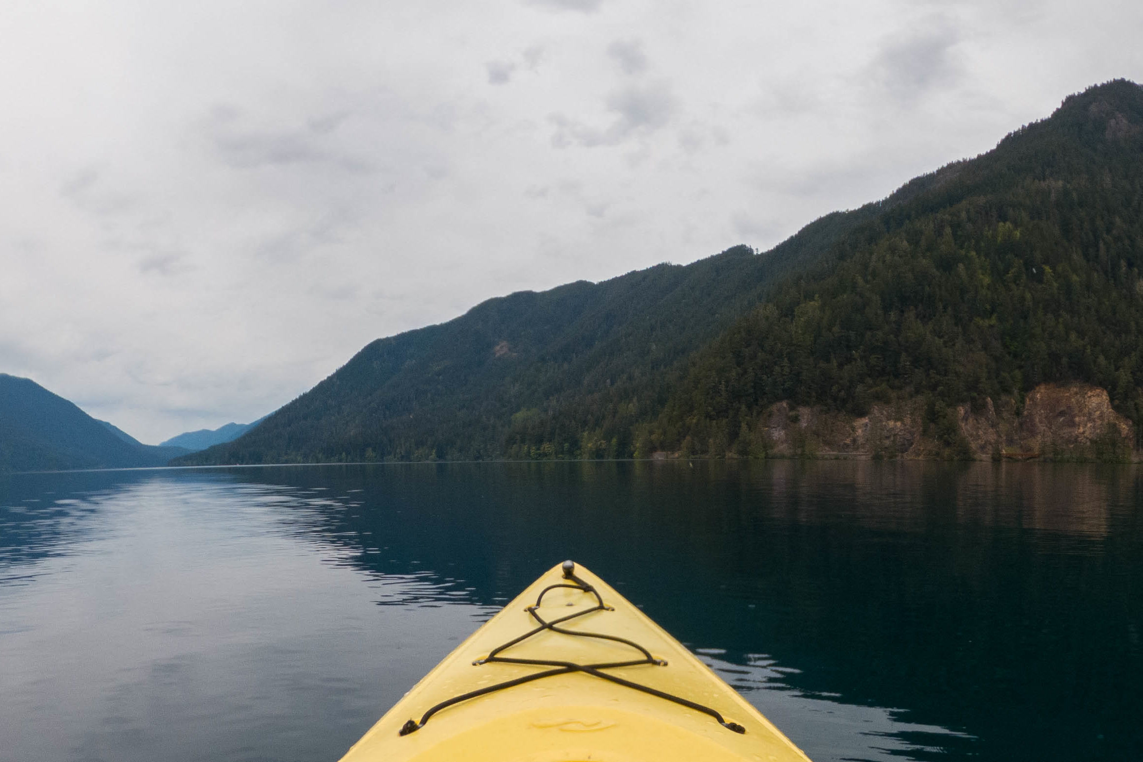 The yellow bow of a kayak pointing across still, dark Lake Crescent toward steep forested mountains under grey clouds