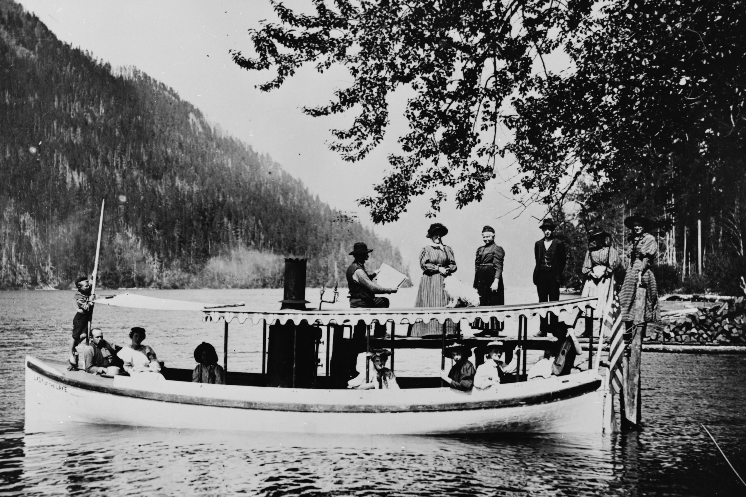 Black-and-white historic photo of passengers in long dresses and hats aboard the Lady of the Lake ferry on Lake Crescent, with forested hills behind