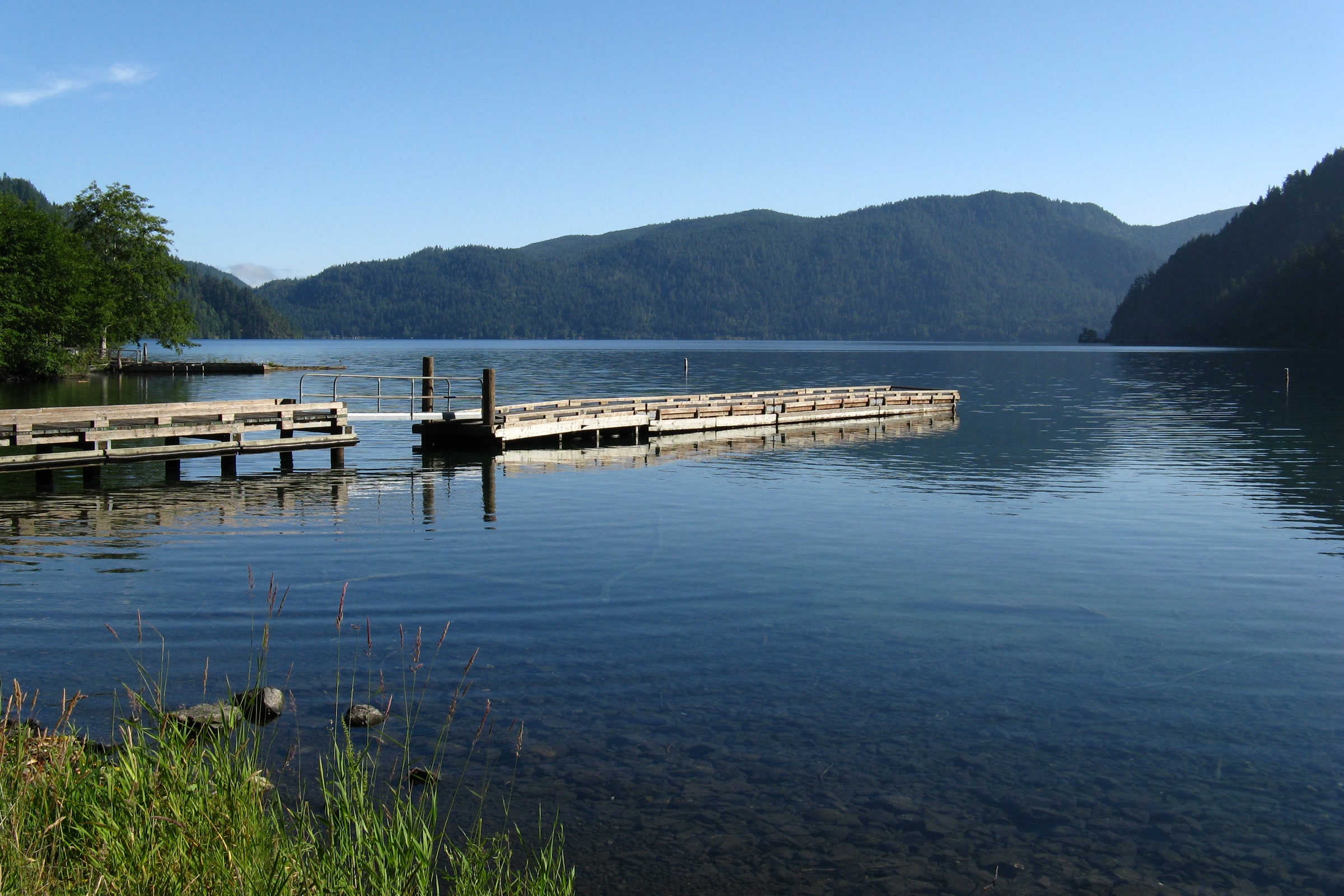 Wooden boat docks reaching into still Lake Crescent, with grassy shore and rounded stones in the foreground