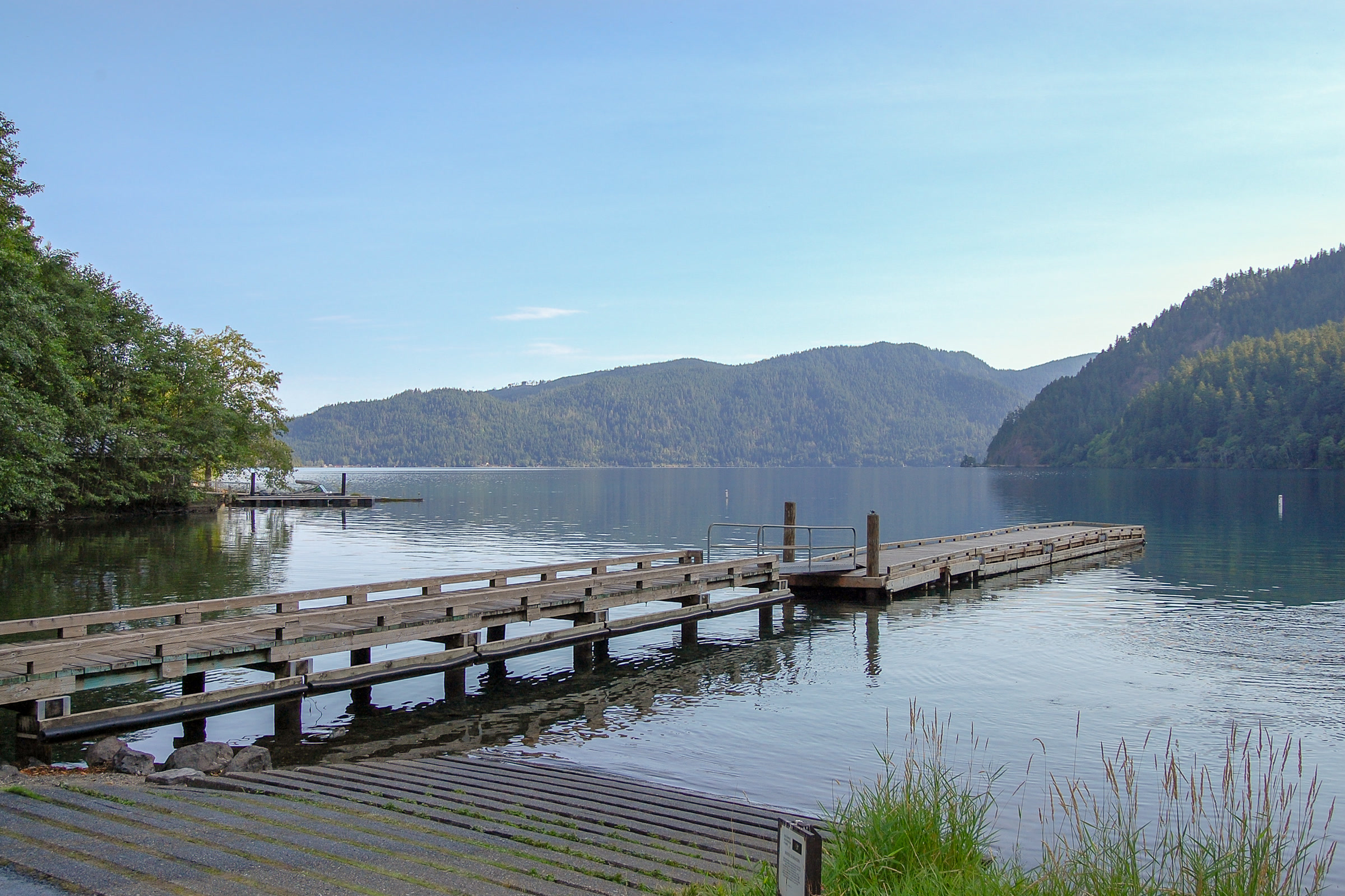 A wooden dock and concrete boat ramp on calm Lake Crescent on a clear morning, with forested mountains across the water