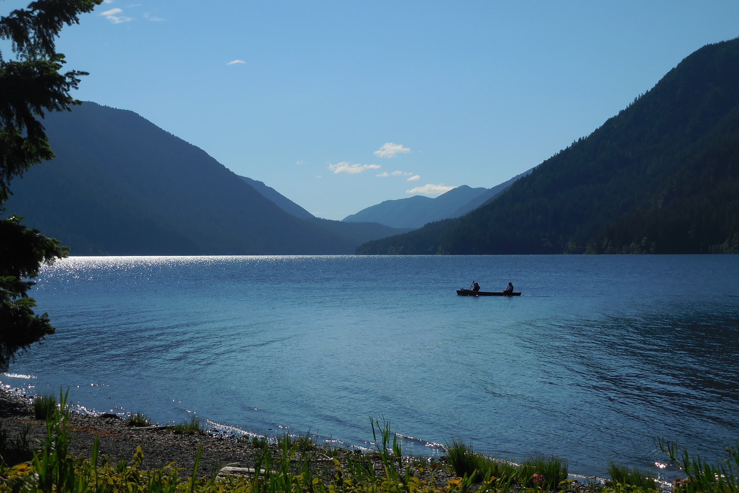 Two paddlers in a canoe on calm blue Lake Crescent, framed by forested mountains and a pebbly shore