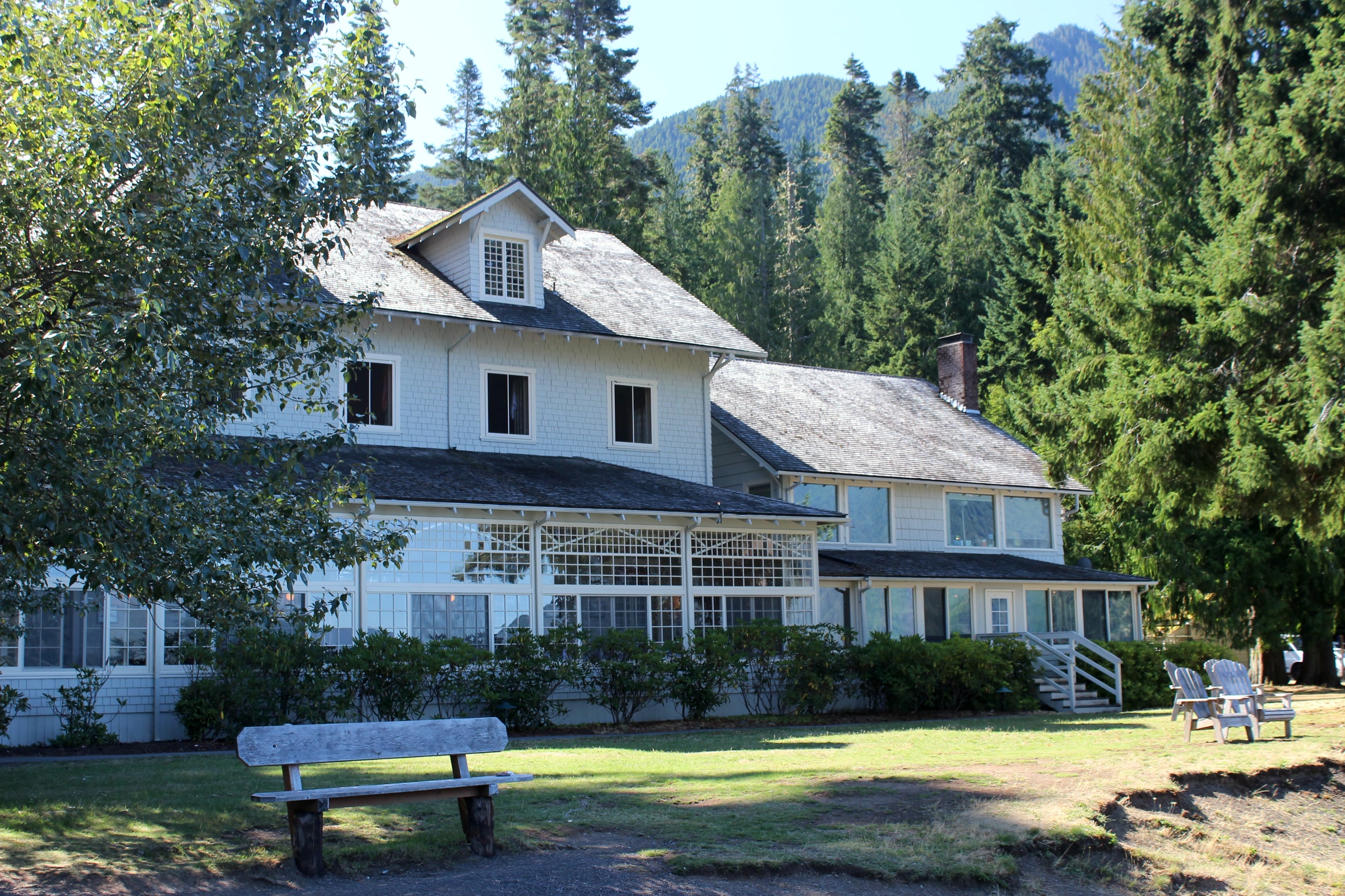 White two-story Lake Crescent Lodge with its glassed-in sun porch and a wooden bench on the lawn, framed by tall firs