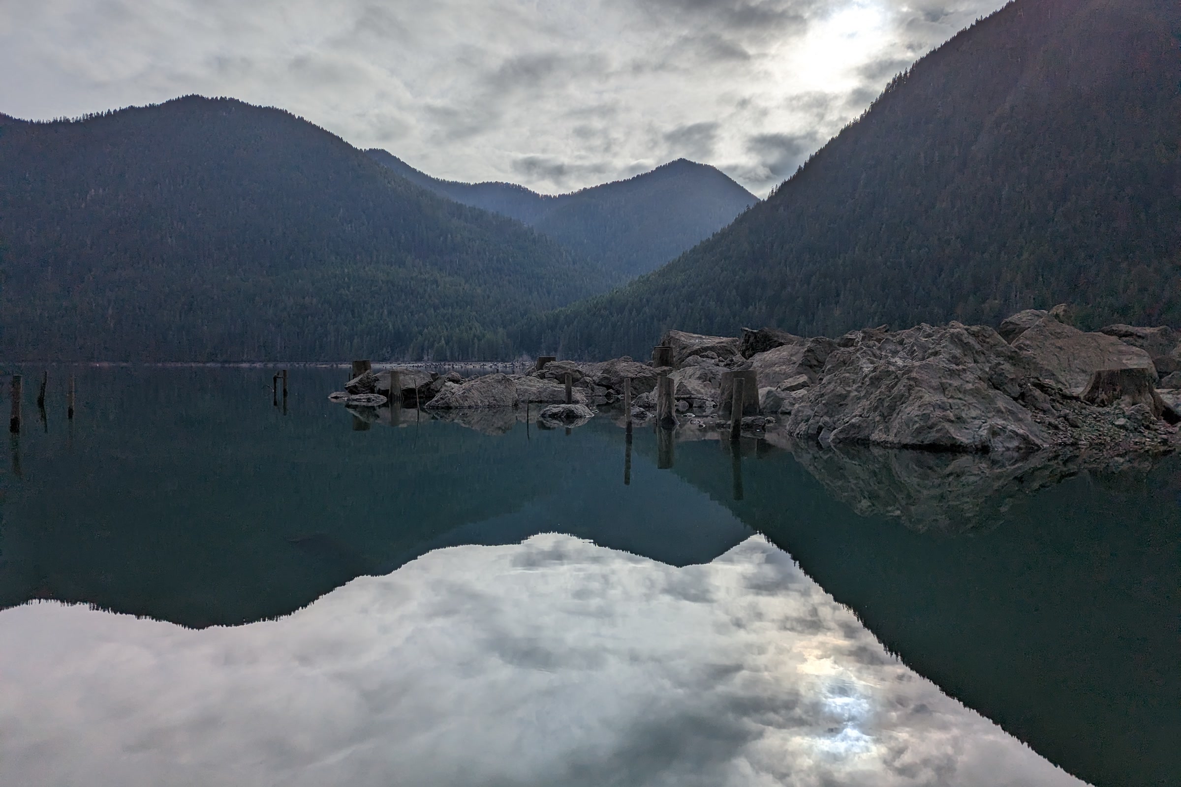 Still water on Lake Cushman reflecting dark forested mountains and a cloudy sky