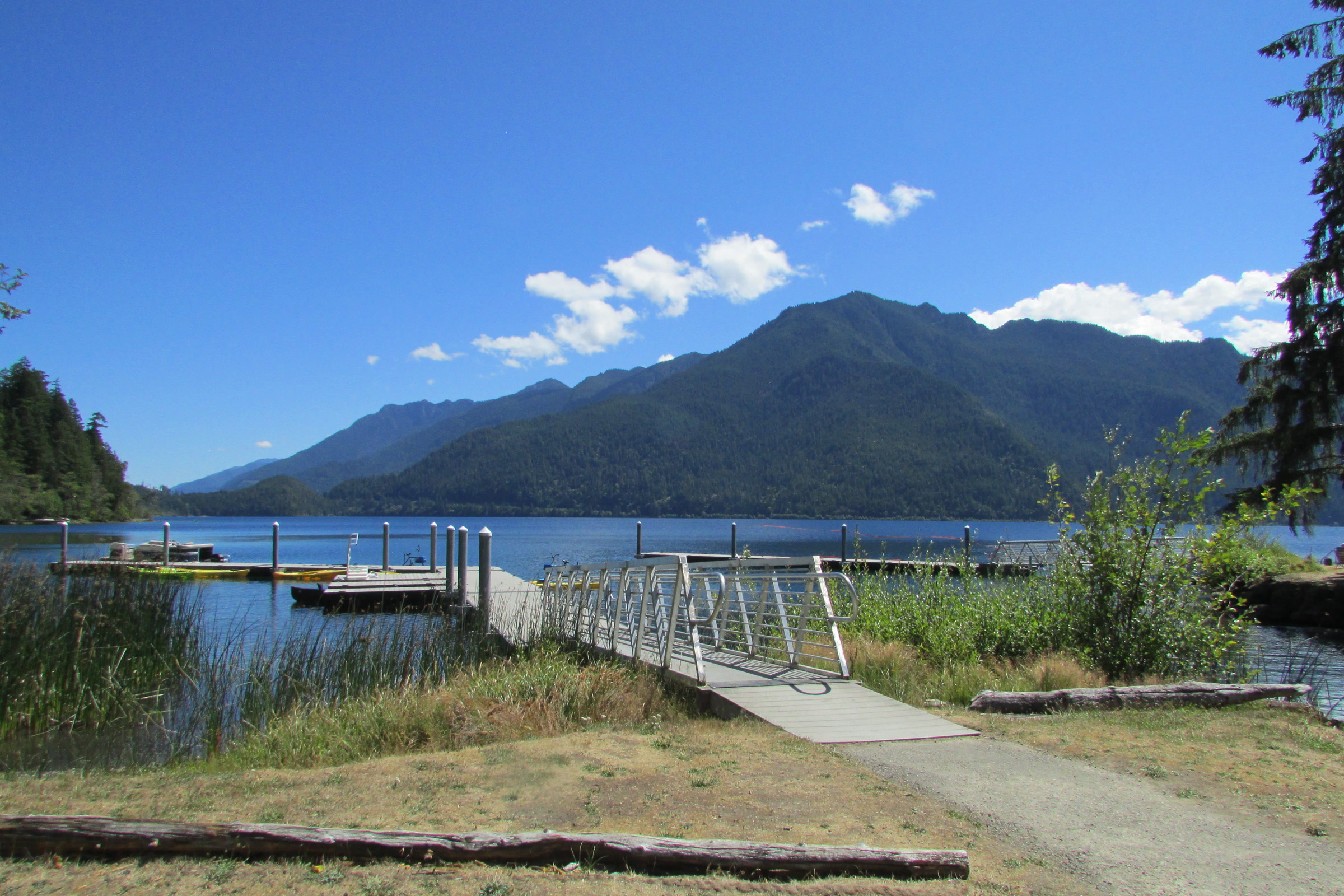 A wooden dock and walkway reaching into Lake Crescent at Log Cabin Resort, with forested mountains and blue sky beyond