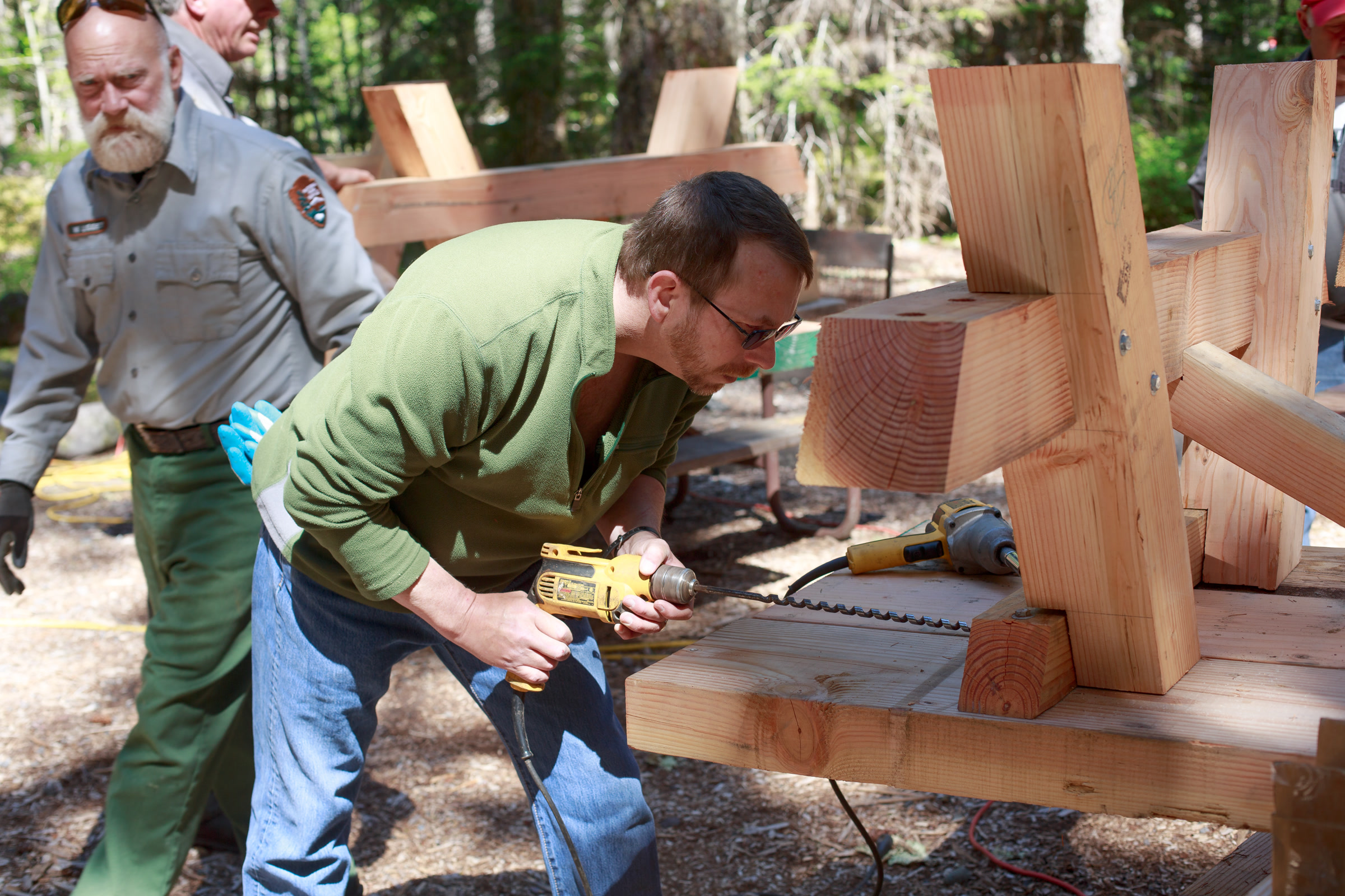 A volunteer drills into a heavy timber frame while a uniformed park employee walks past at a forest work site