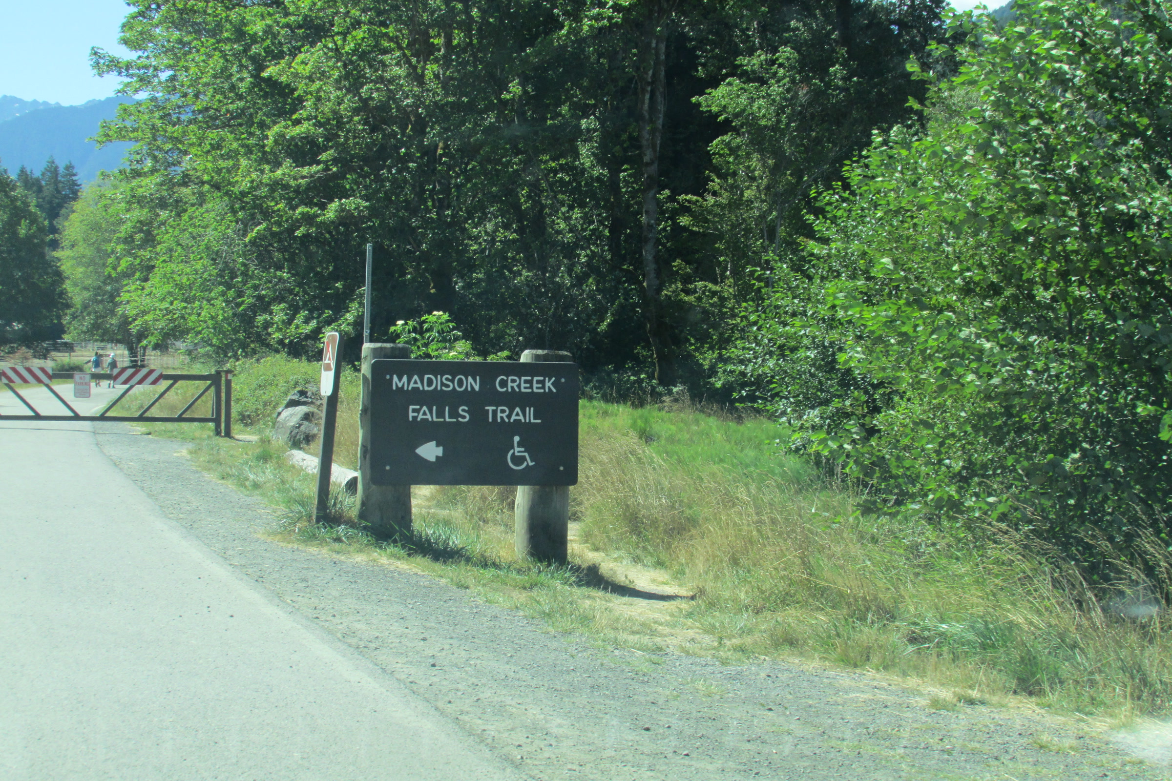 A Madison Creek Falls Trail sign with a wheelchair symbol beside the paved road and a closed gate in the Elwha Valley