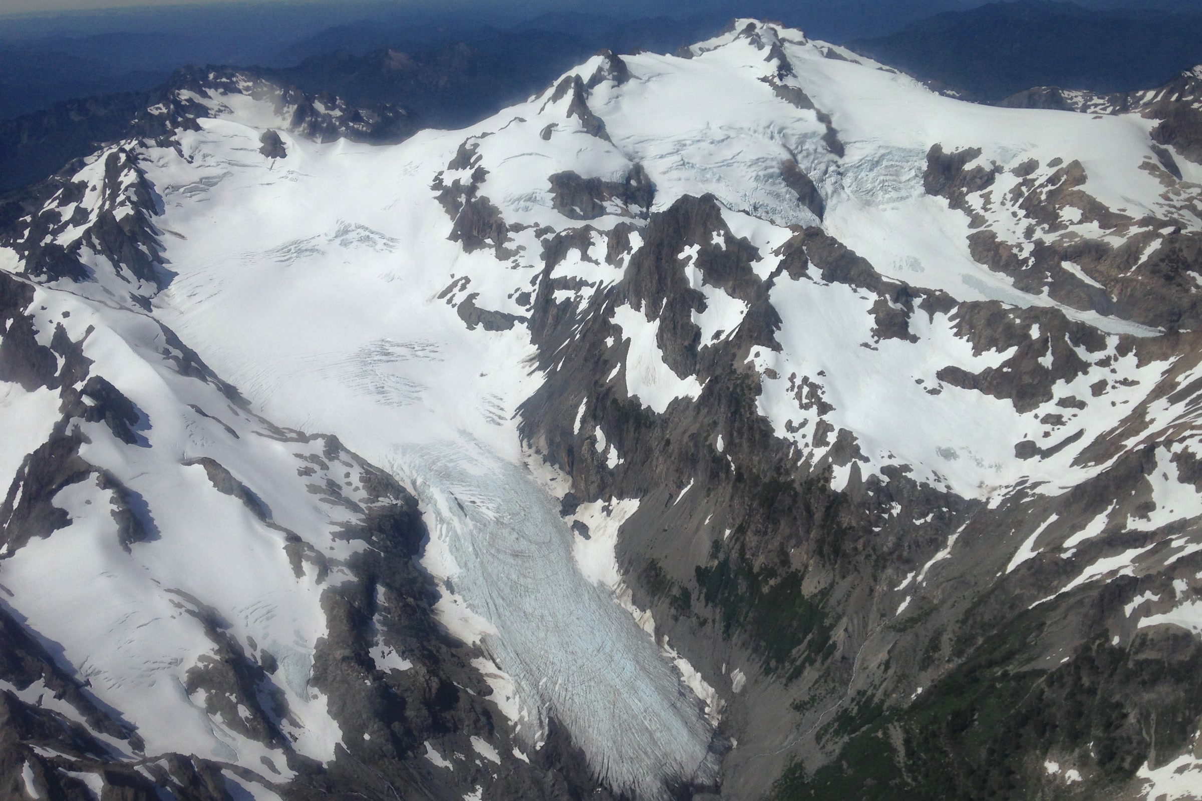 Aerial view of Mount Olympus in July with snowfields and a broad glacier tongue flowing down between dark rock ridges