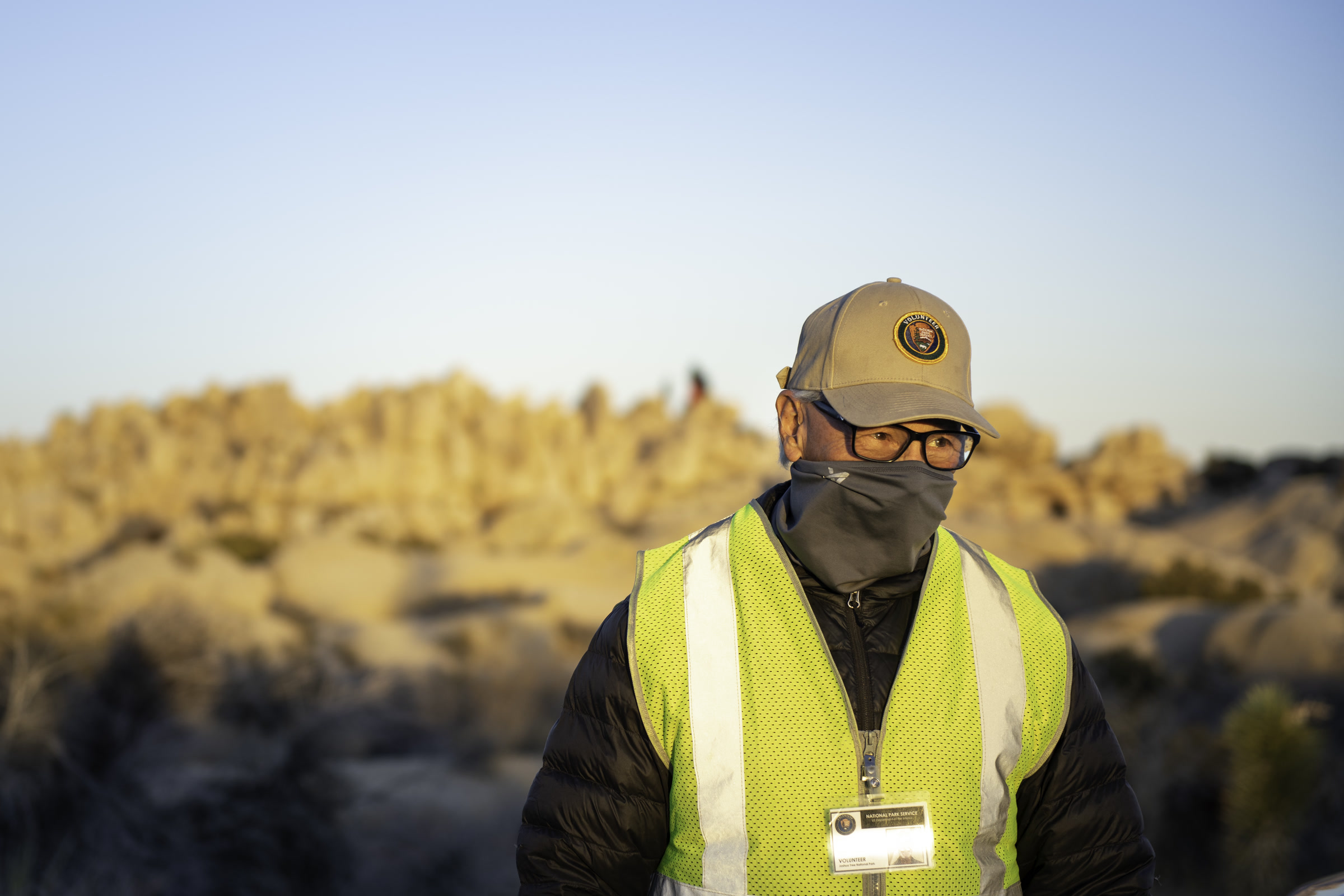 A national park volunteer in a tan volunteer cap, face mask and yellow safety vest with an ID badge, desert rocks glowing behind