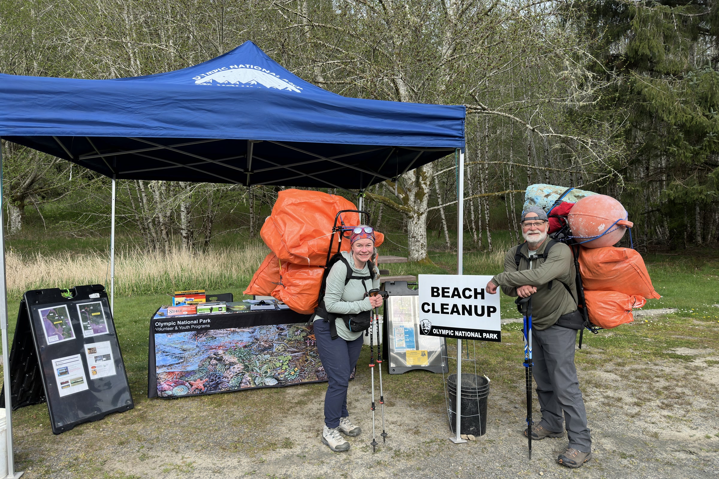 Two coastal cleanup volunteers with backpacks loaded with orange debris bags and a buoy beside an Olympic National Park beach cleanup sign