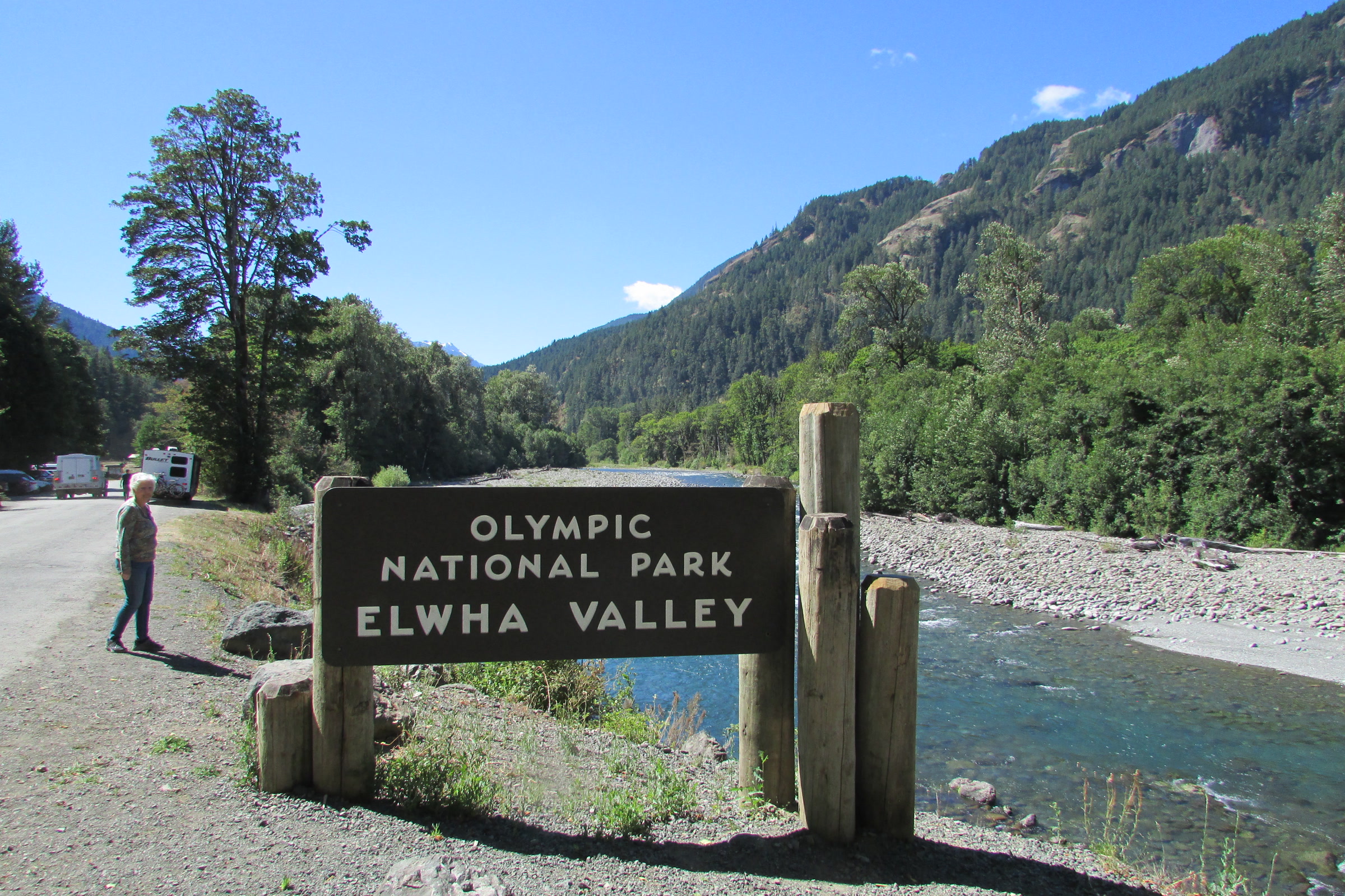Wooden Olympic National Park Elwha Valley sign beside the gravel bar of the Elwha River, with forested mountains under a blue sky