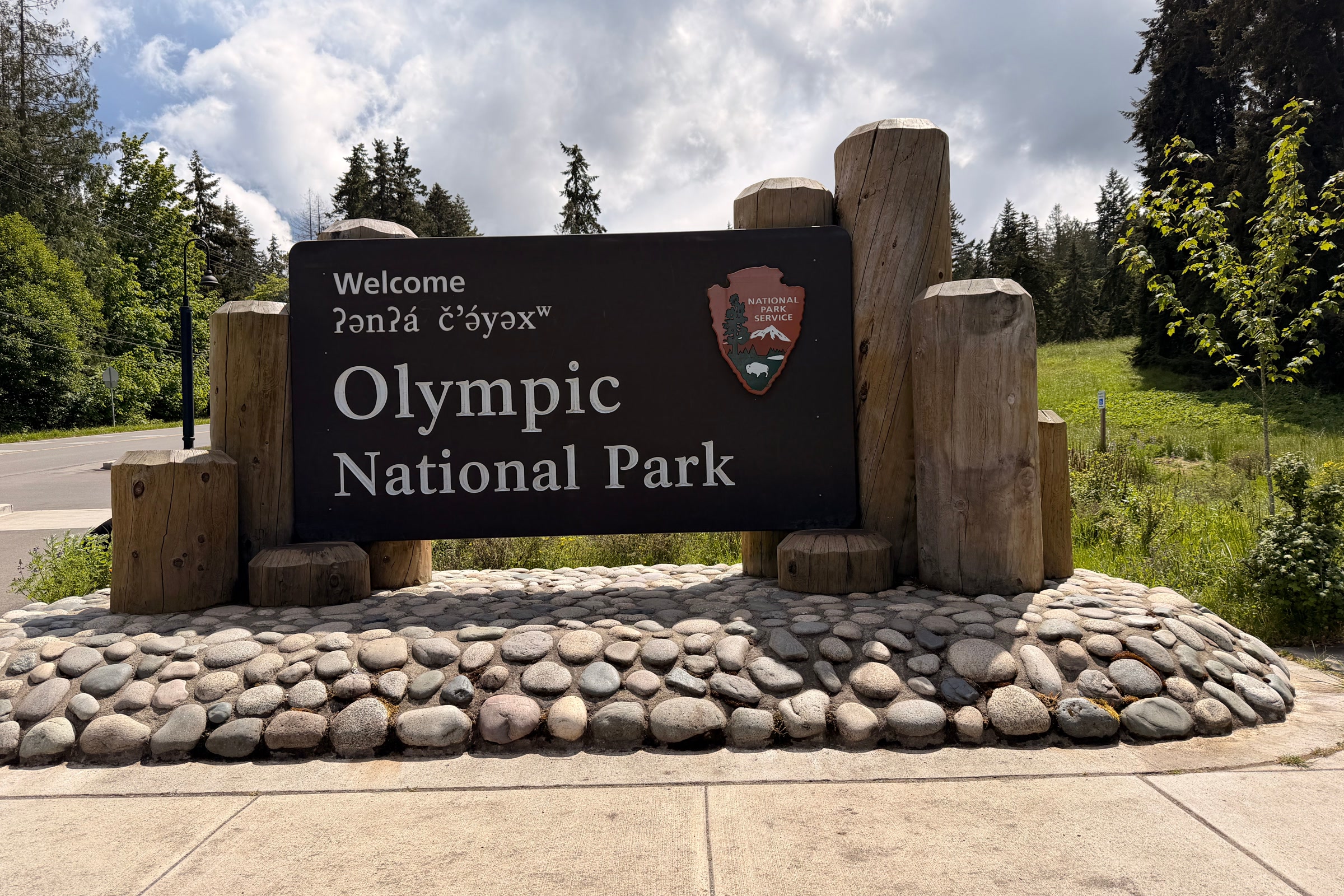 Olympic National Park welcome sign with a Klallam greeting, framed by cedar posts on a river-cobble base with grass and young trees behind