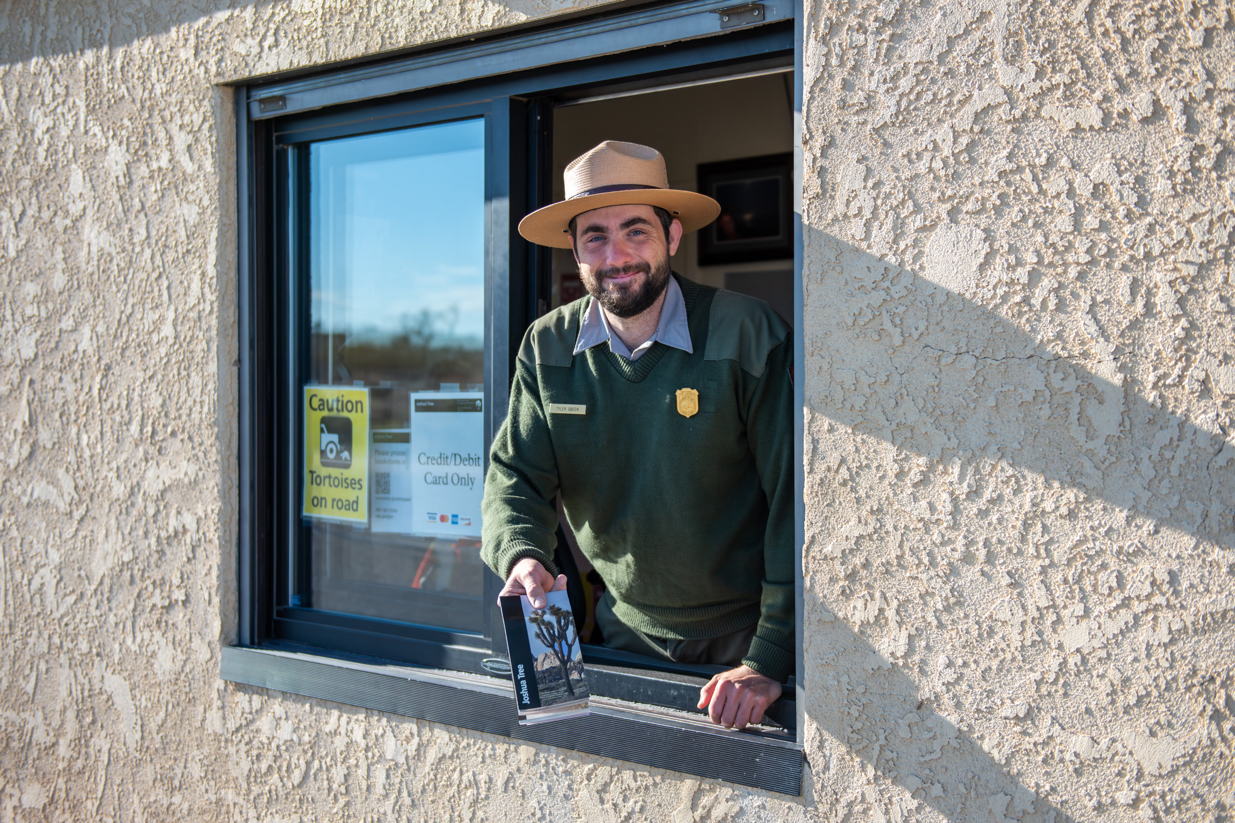 A smiling park ranger in a flat hat leans out of an entrance station window holding a park brochure