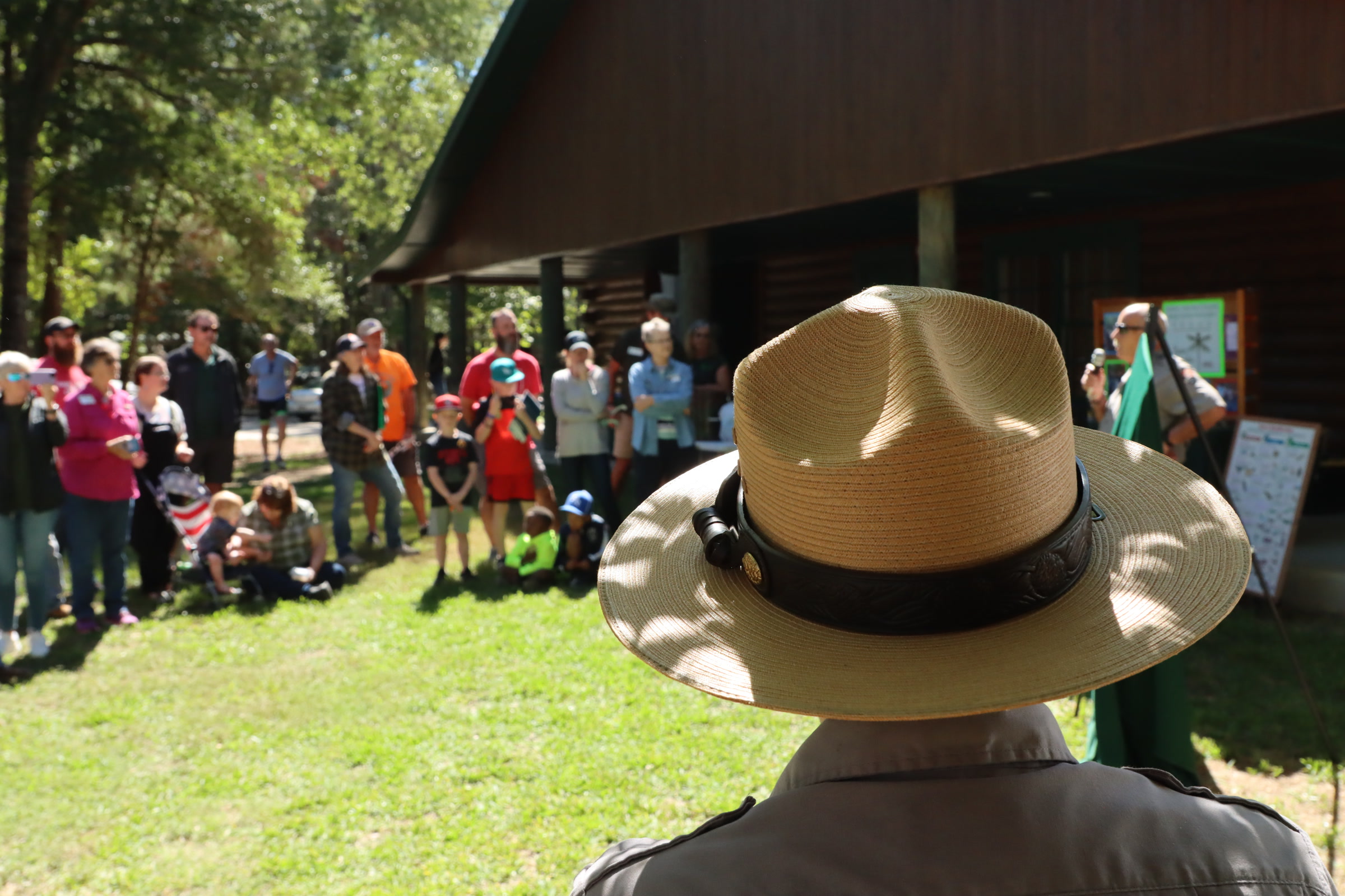 A park ranger in a tan flat hat, seen from behind, watching families gathered outdoors for a ranger program