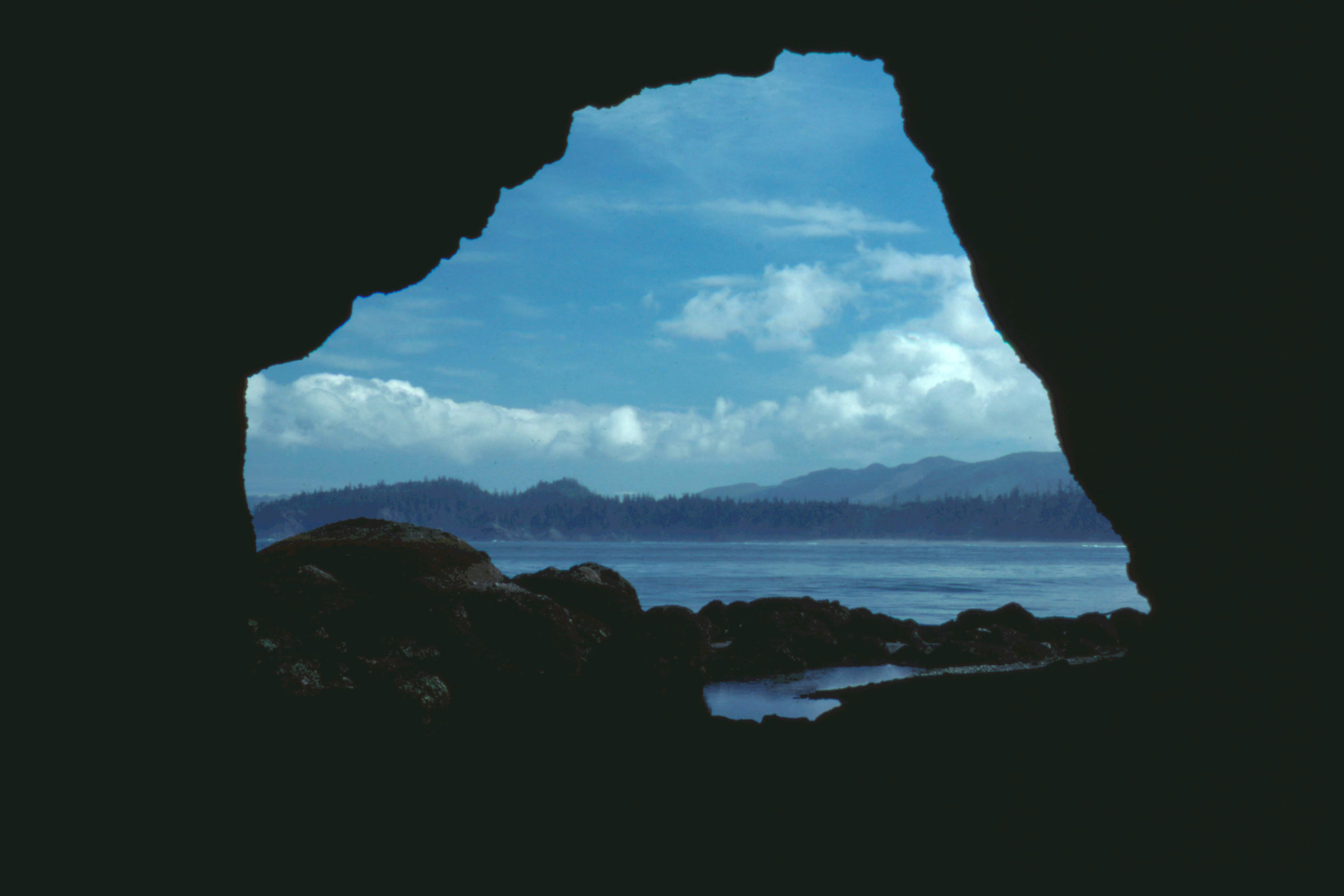 View out through a dark rock arch at Point of Arches to blue sky, white clouds, calm water and a distant forested shore
