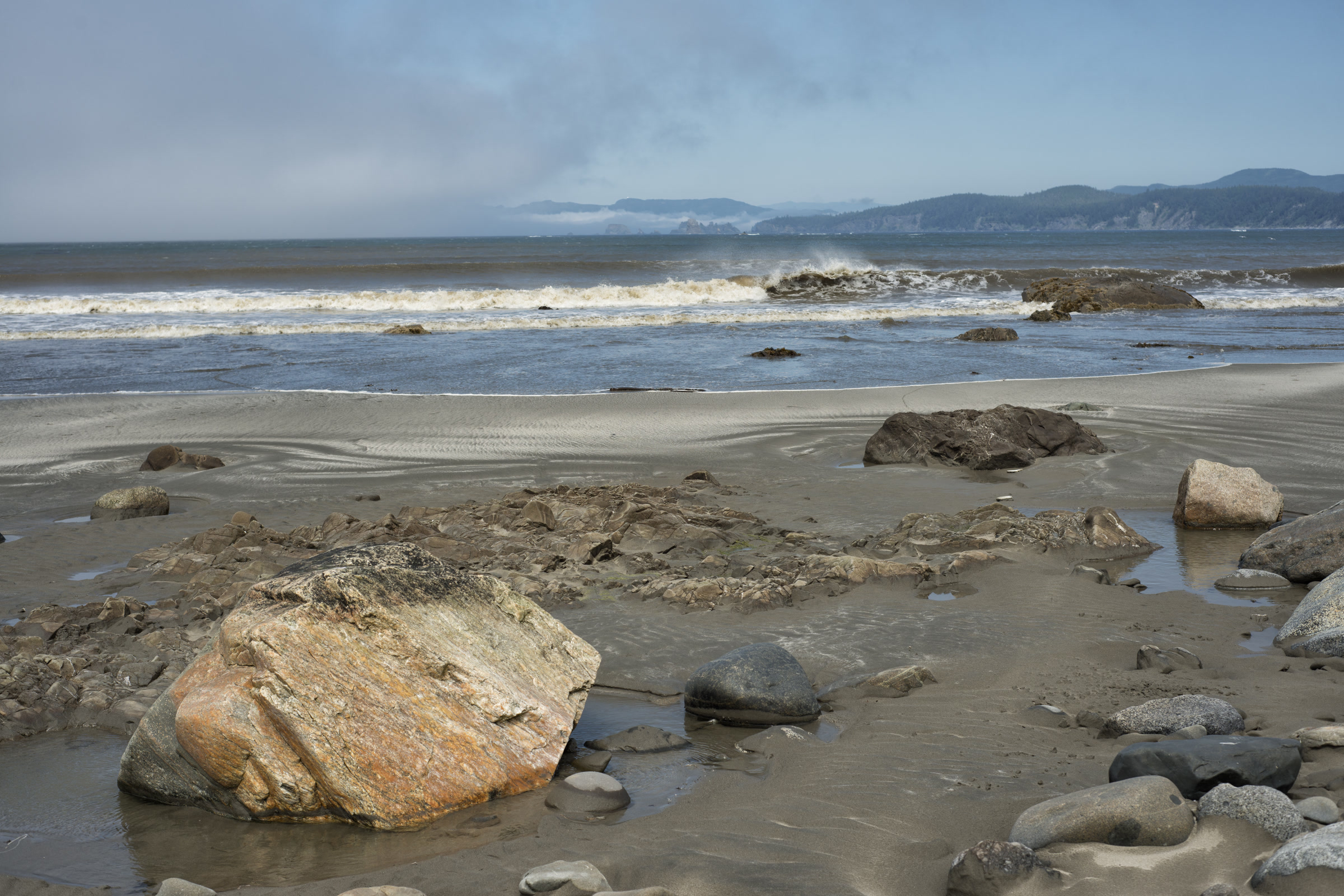 A sandy beach scattered with boulders and flat rock ledges, with surf and the distant sea stacks of Point of Arches under low cloud