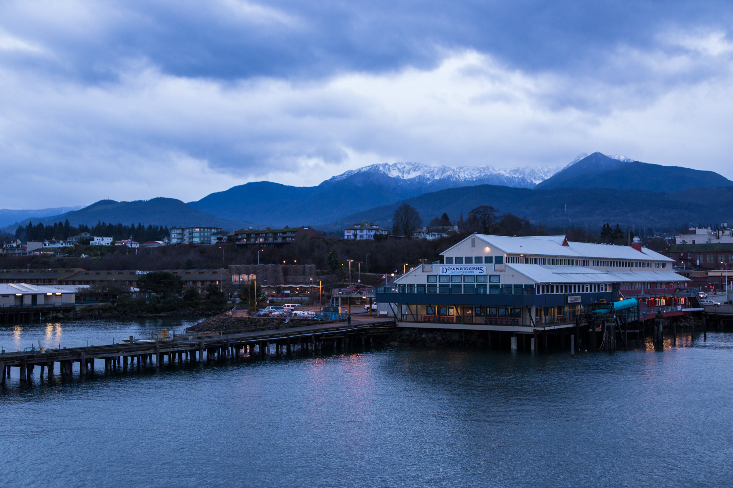 Port Angeles waterfront and pier buildings at dusk in January, with snow covered Olympic Mountains under grey clouds behind the town