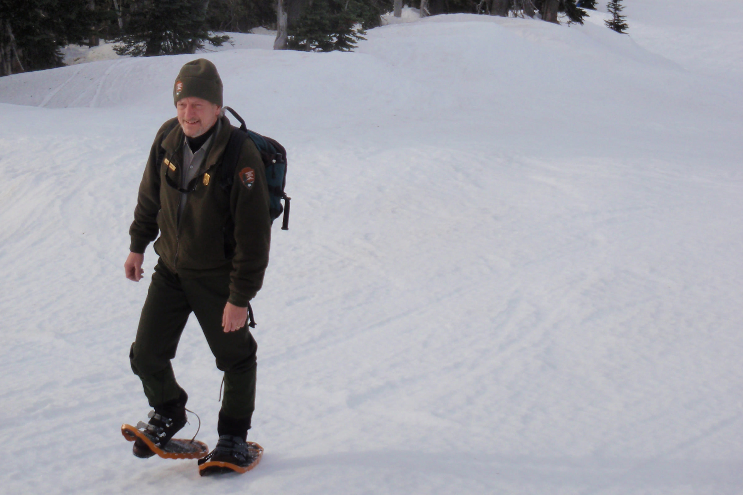 A park ranger in uniform and wool hat walking across deep snow on snowshoes at Hurricane Ridge