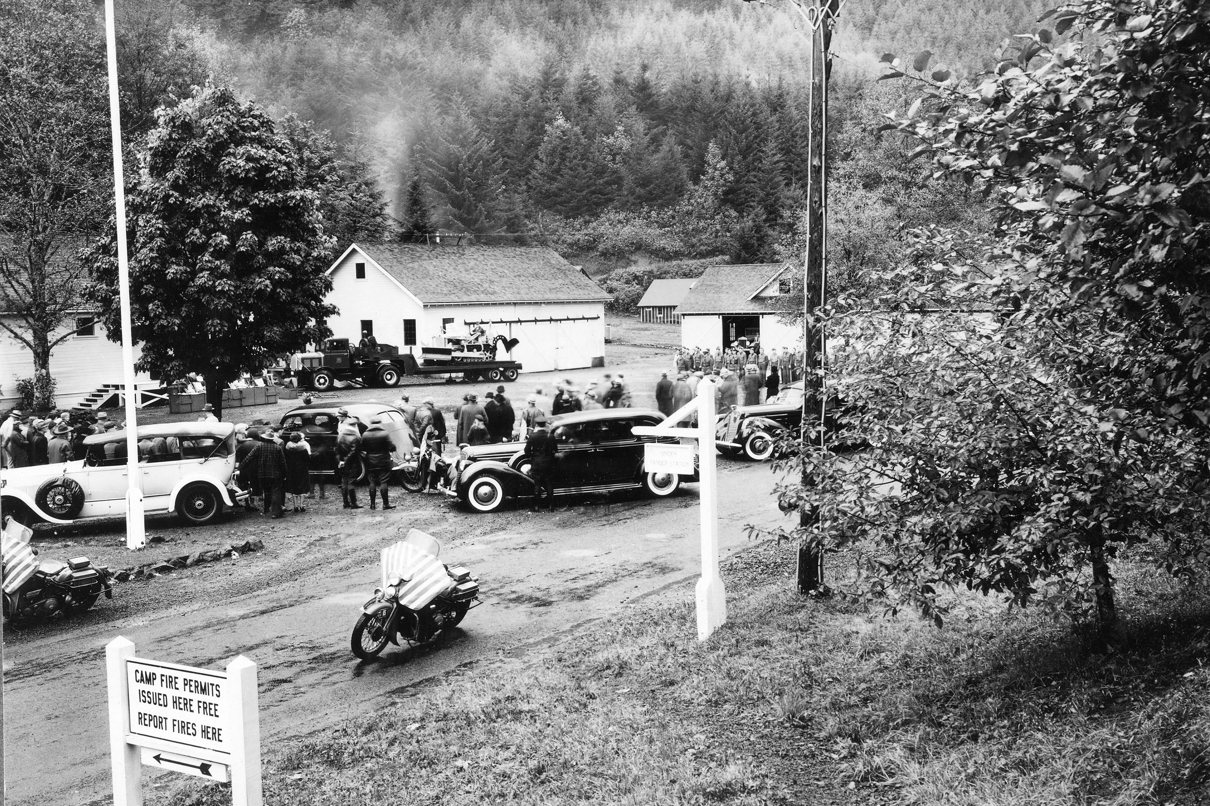 Black and white photo of President Roosevelt's motorcade and a crowd gathered by white ranger station buildings in a forest clearing