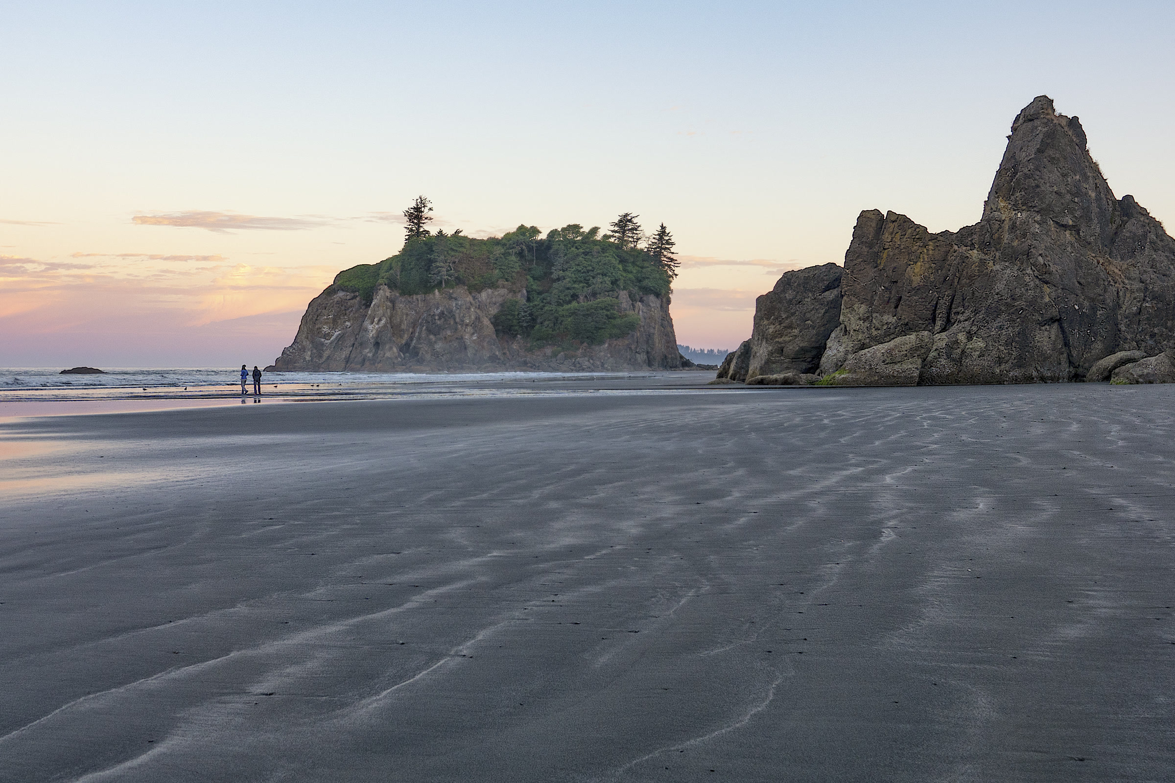 Abbey Island and a jagged sea stack at Ruby Beach in soft evening light, with ripples in the wet sand and two small figures walking