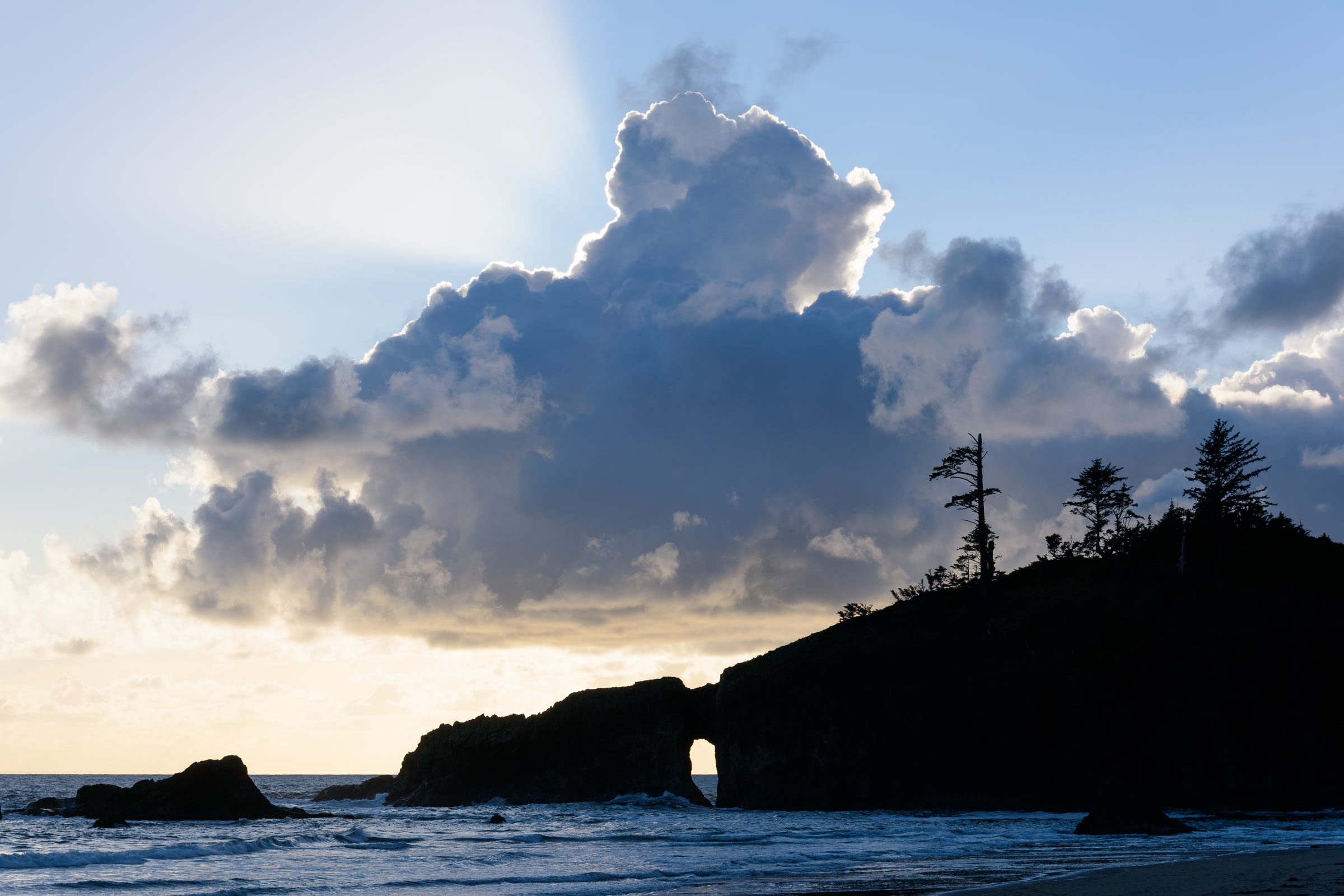 The dark silhouette of a natural rock arch and forested headland at Second Beach under a towering cloud at sunset