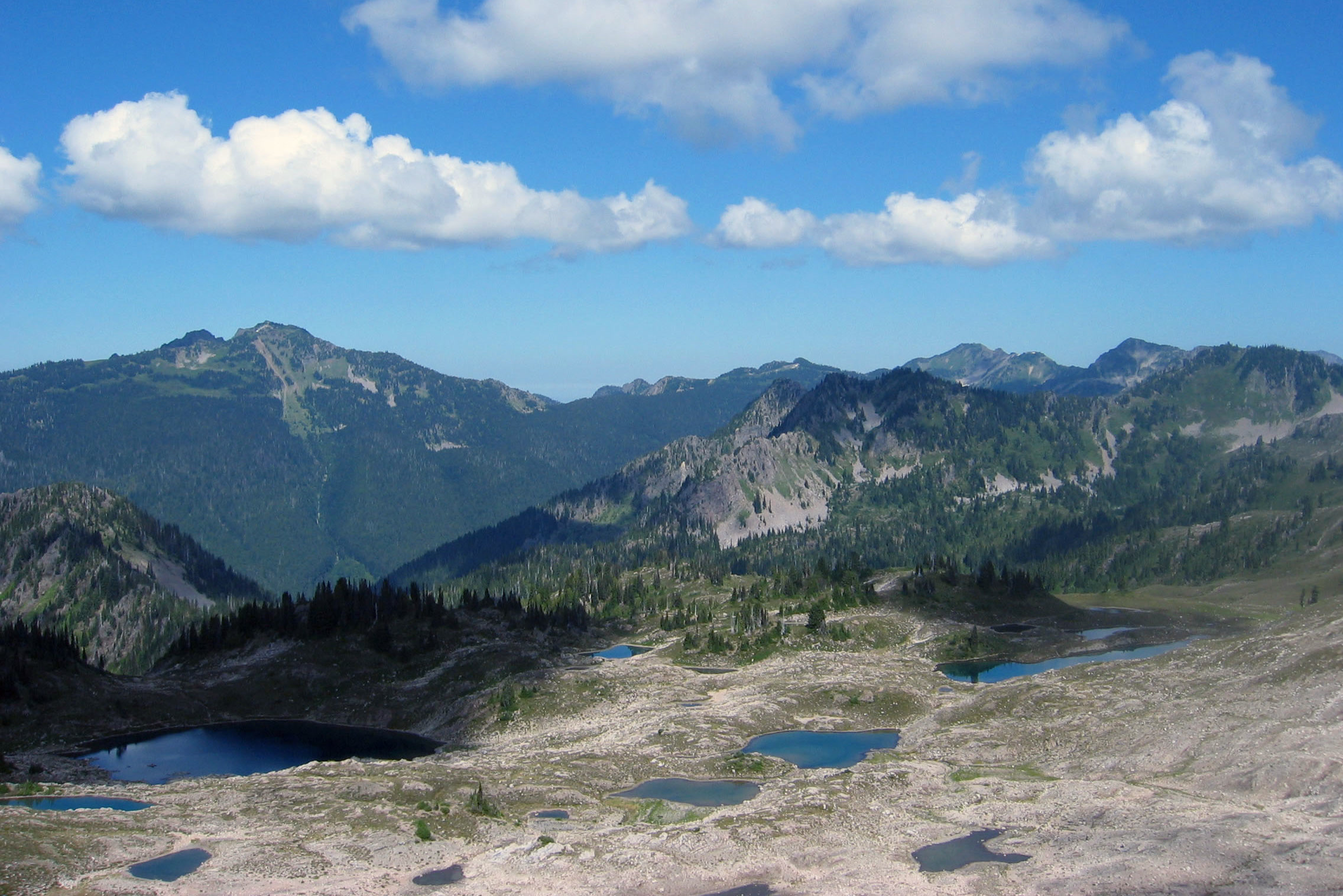 Several small blue lakes in the open, rocky floor of Seven Lakes Basin, with green forested mountains and white clouds beyond