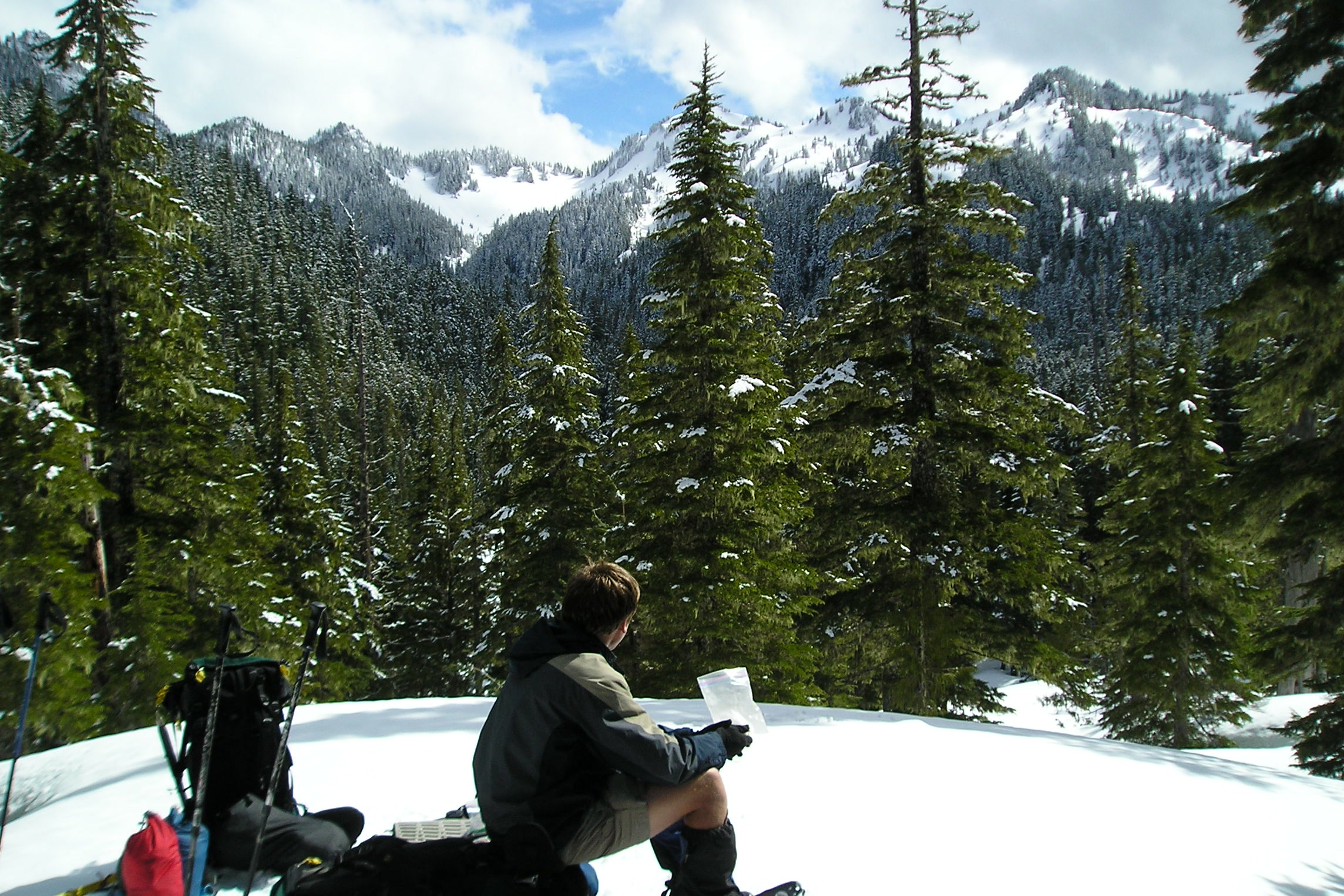 A snowshoer sitting on a snowbank for lunch beside backpacks, with snowy forested peaks behind