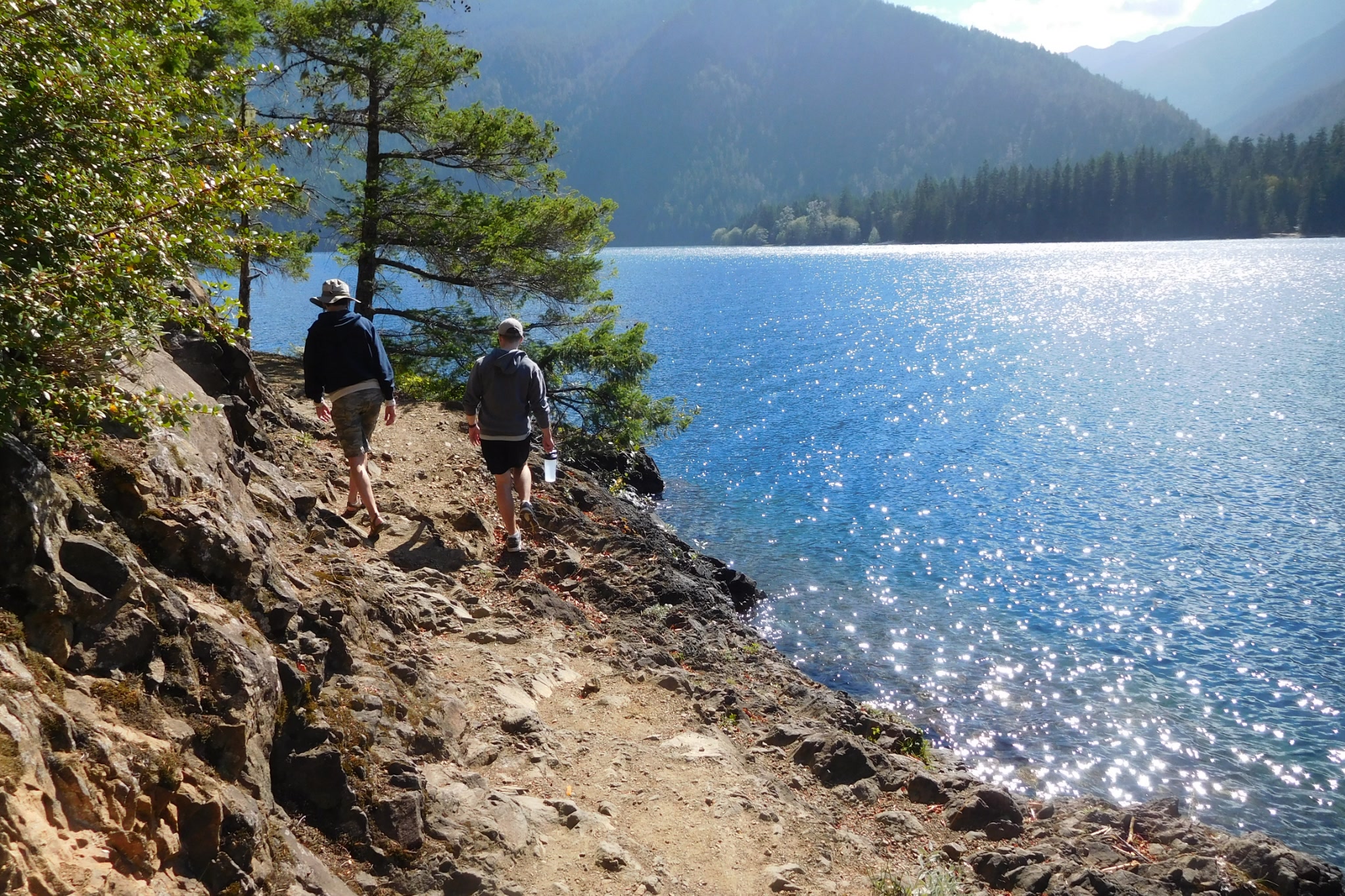 Two walkers on a rocky shoreline path beside sparkling blue Lake Crescent, with forested mountains across the water